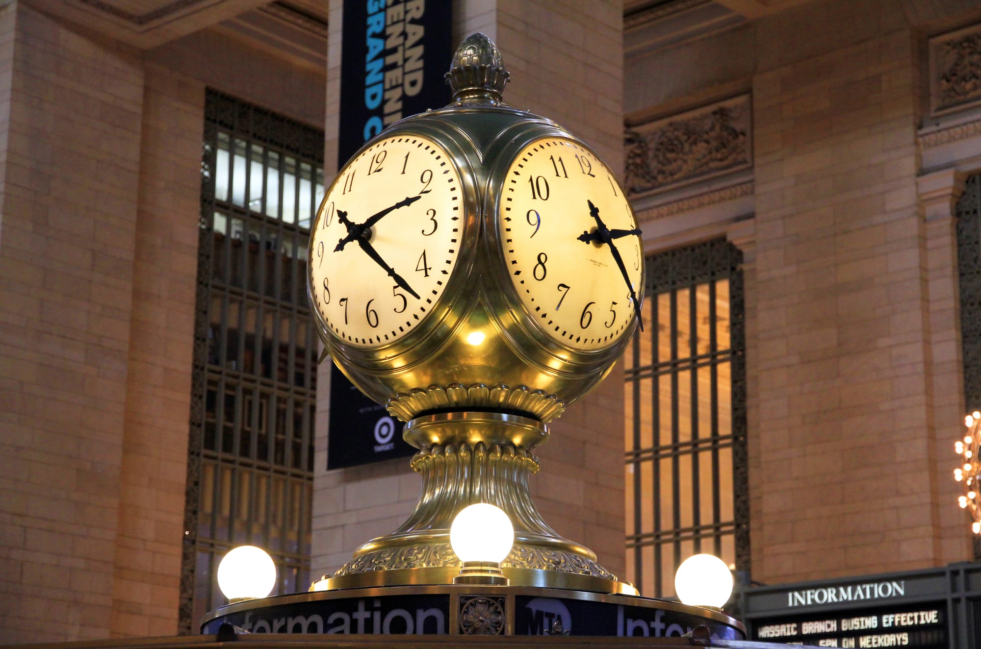 A brass four-faced clock with glowing globe lamps on top of an information booth in a large stone hall