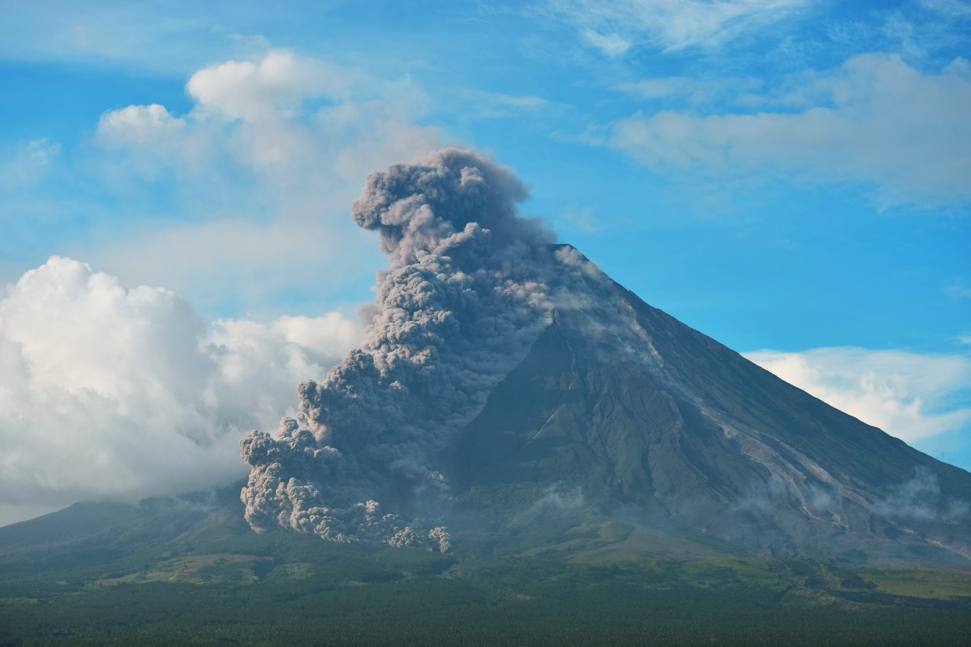 A steep, symmetrical cone-shaped mountain under a blue sky, with a grey column of ash billowing down one flank