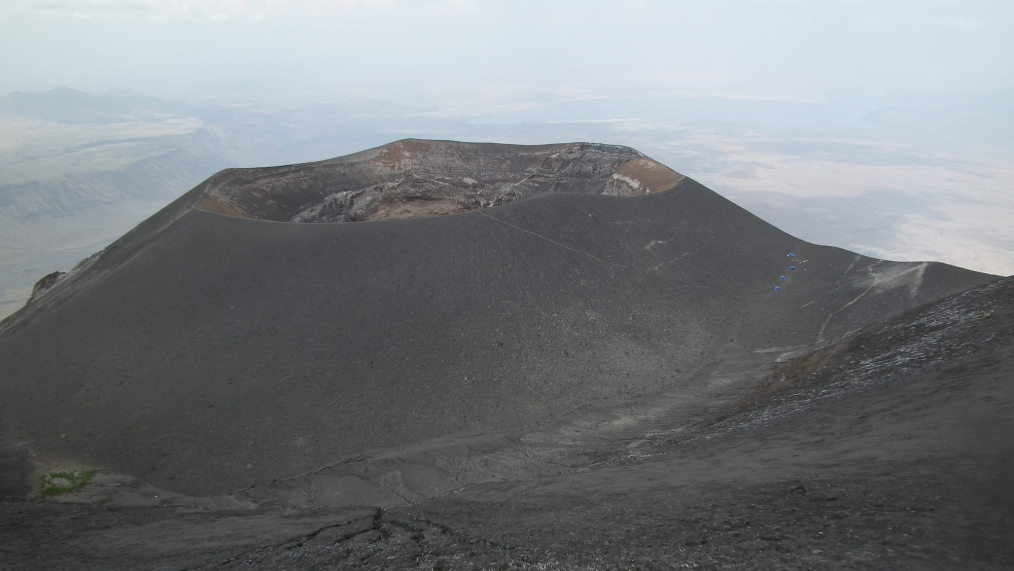 The grey, ash-covered crater rim of a volcano seen from its upper slopes, with a hazy plain stretching away below