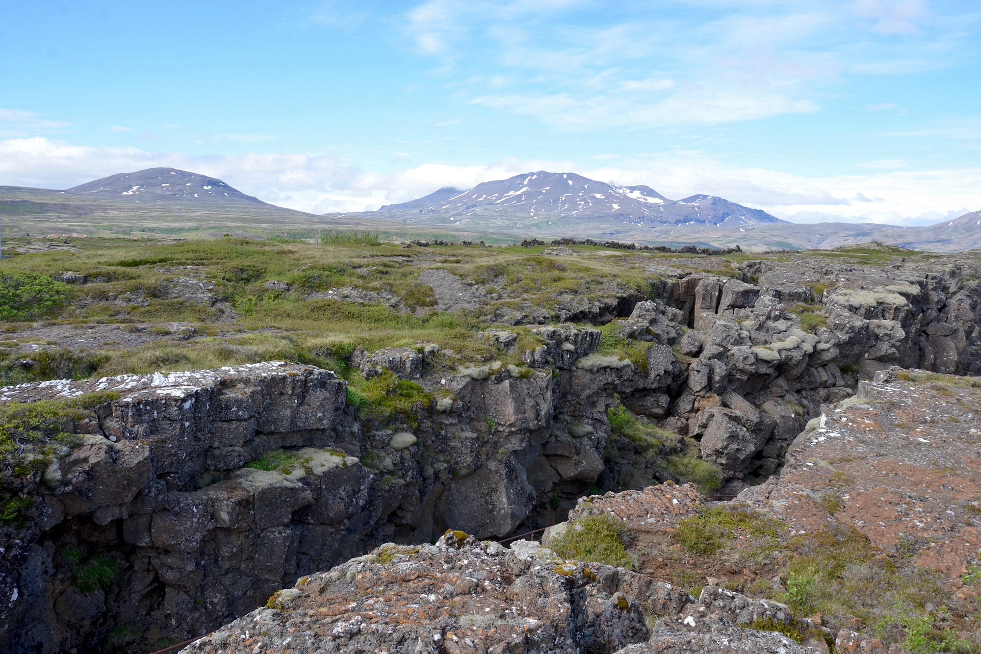 A deep crack in dark, mossy volcanic rock running across a grassy plain towards distant snow-streaked mountains