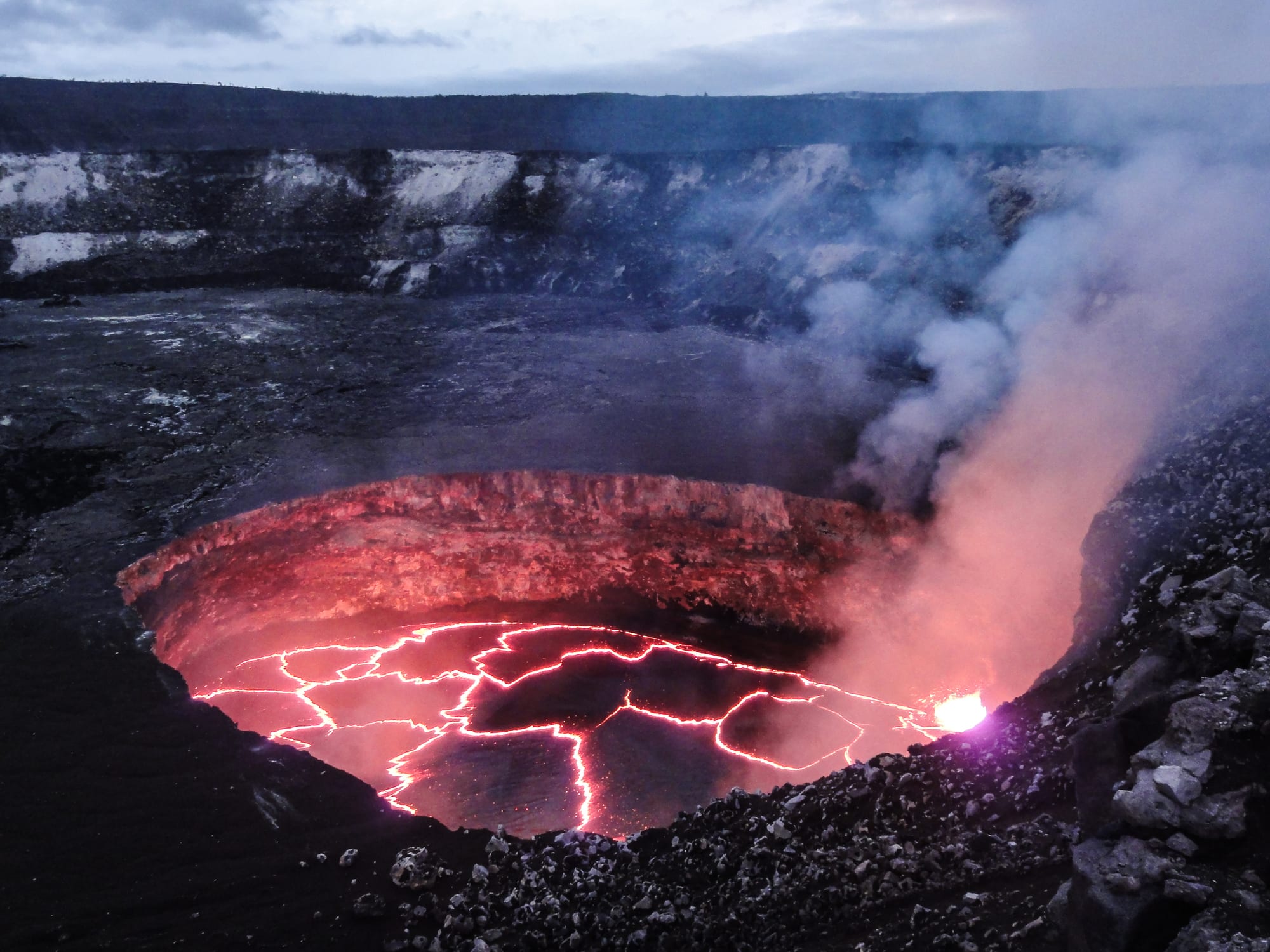 A glowing lava lake with a crust cracked by red lines inside a steep-walled crater, with fumes rising at dusk