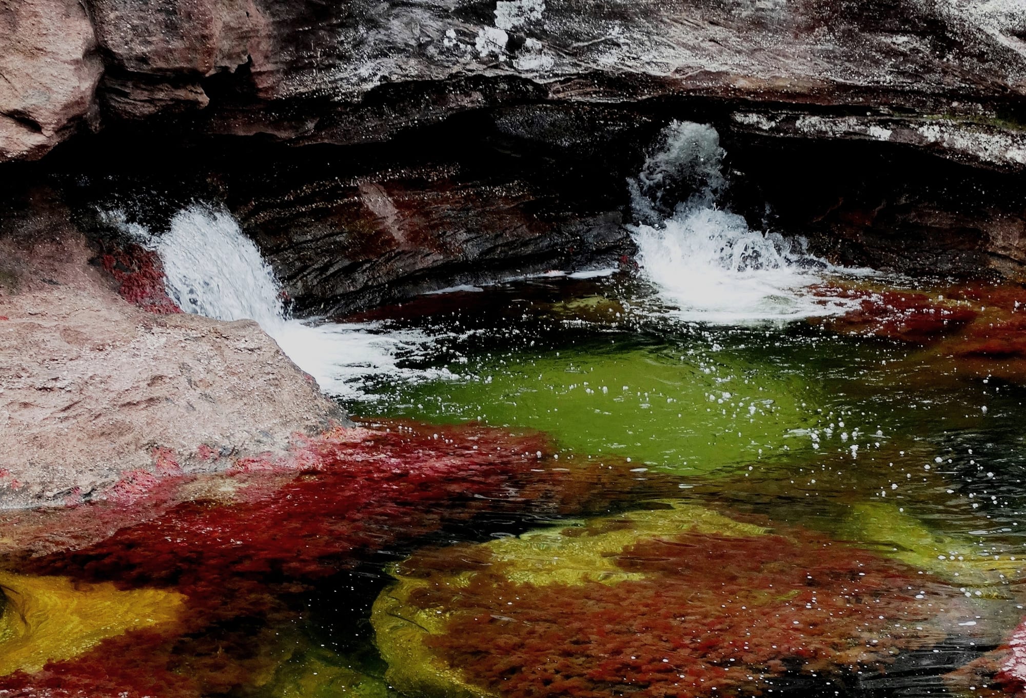 Two small waterfalls tumbling into a clear pool whose bed is patched with deep red, bright green and yellow