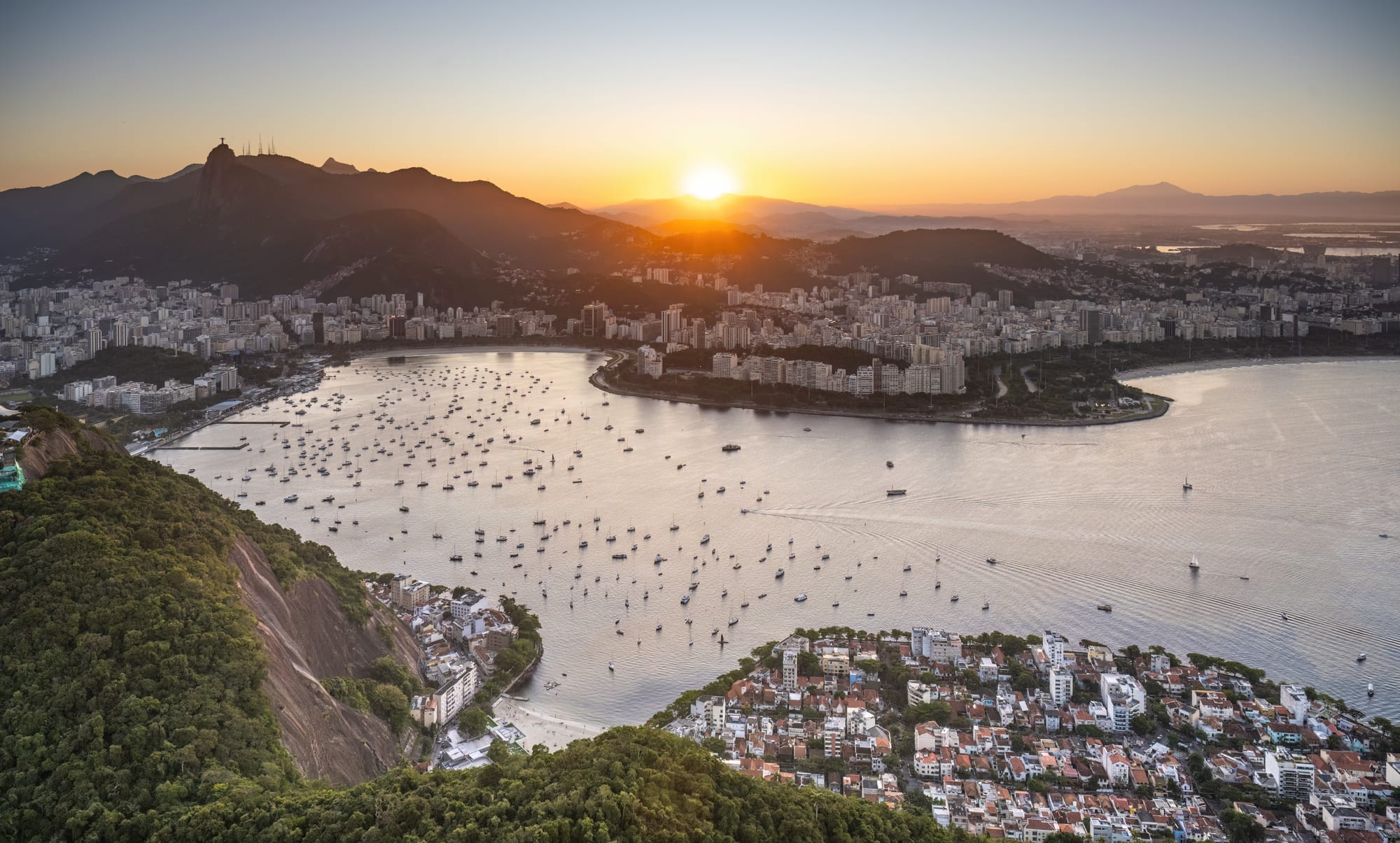 A bay full of moored boats at sunset, with city blocks along the shore and a dark mountain peak topped by a tiny statue on the left