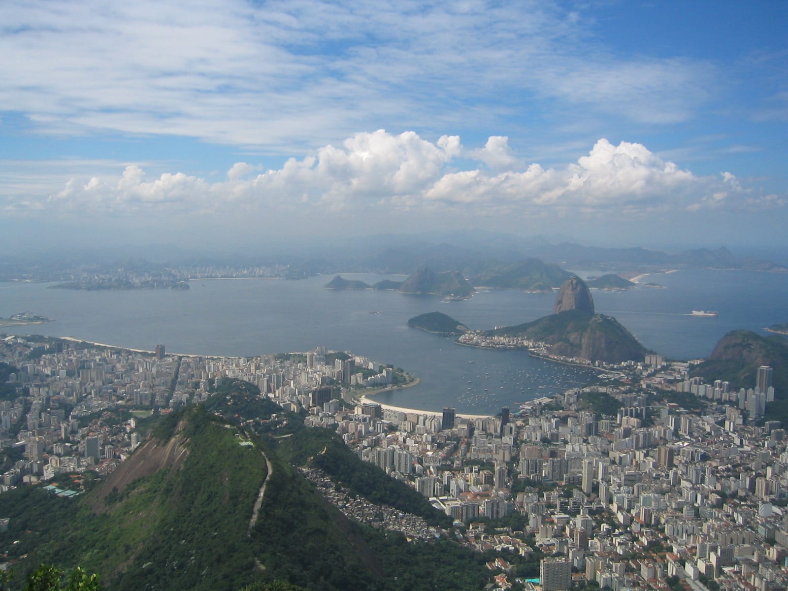 A wide view over a dense city of white towers, a curving beach and a bay with a dome-shaped rock peak, seen from a green mountainside