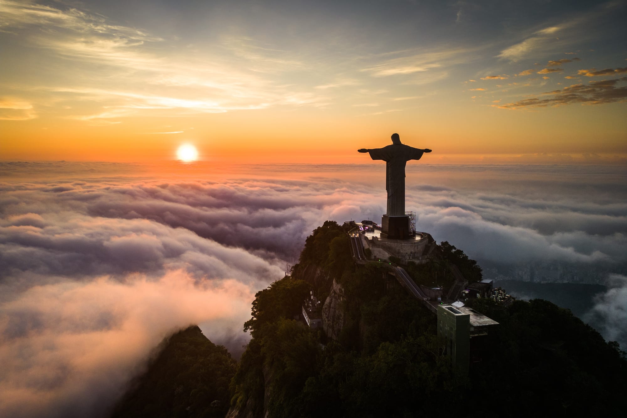 A giant statue of Christ with outstretched arms in silhouette on a forested peak, rising above a sea of clouds at sunrise