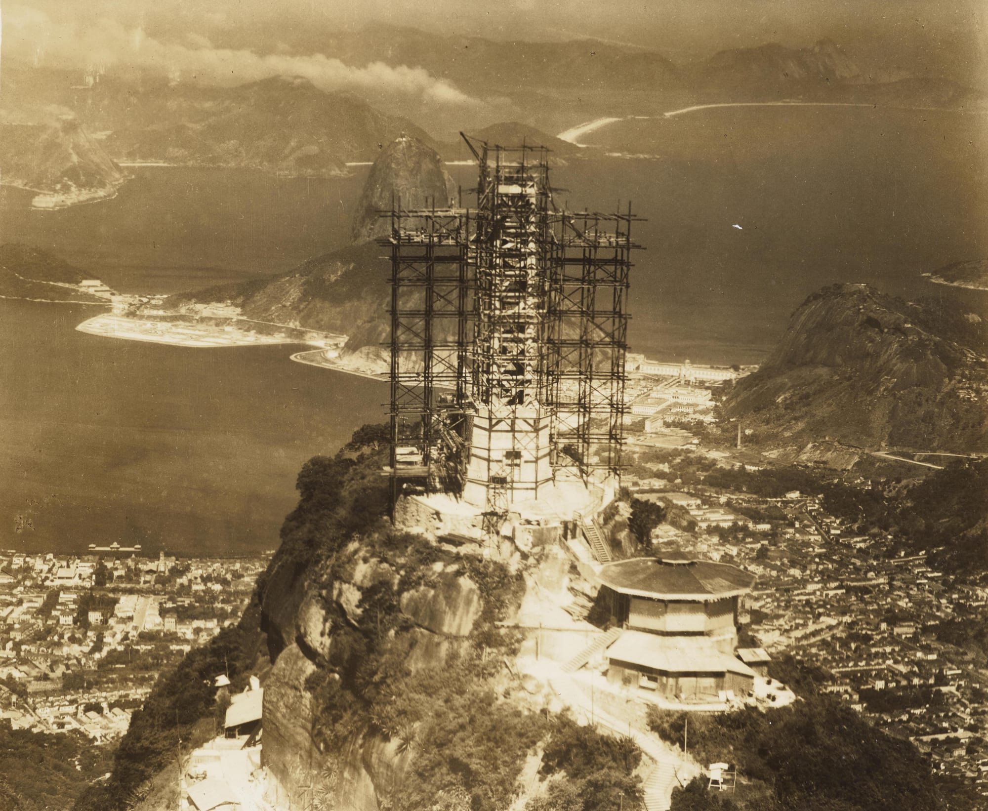 An old sepia photograph of a tall statue wrapped in scaffolding on a rocky summit, with a bay, a domed rock peak and a city far below
