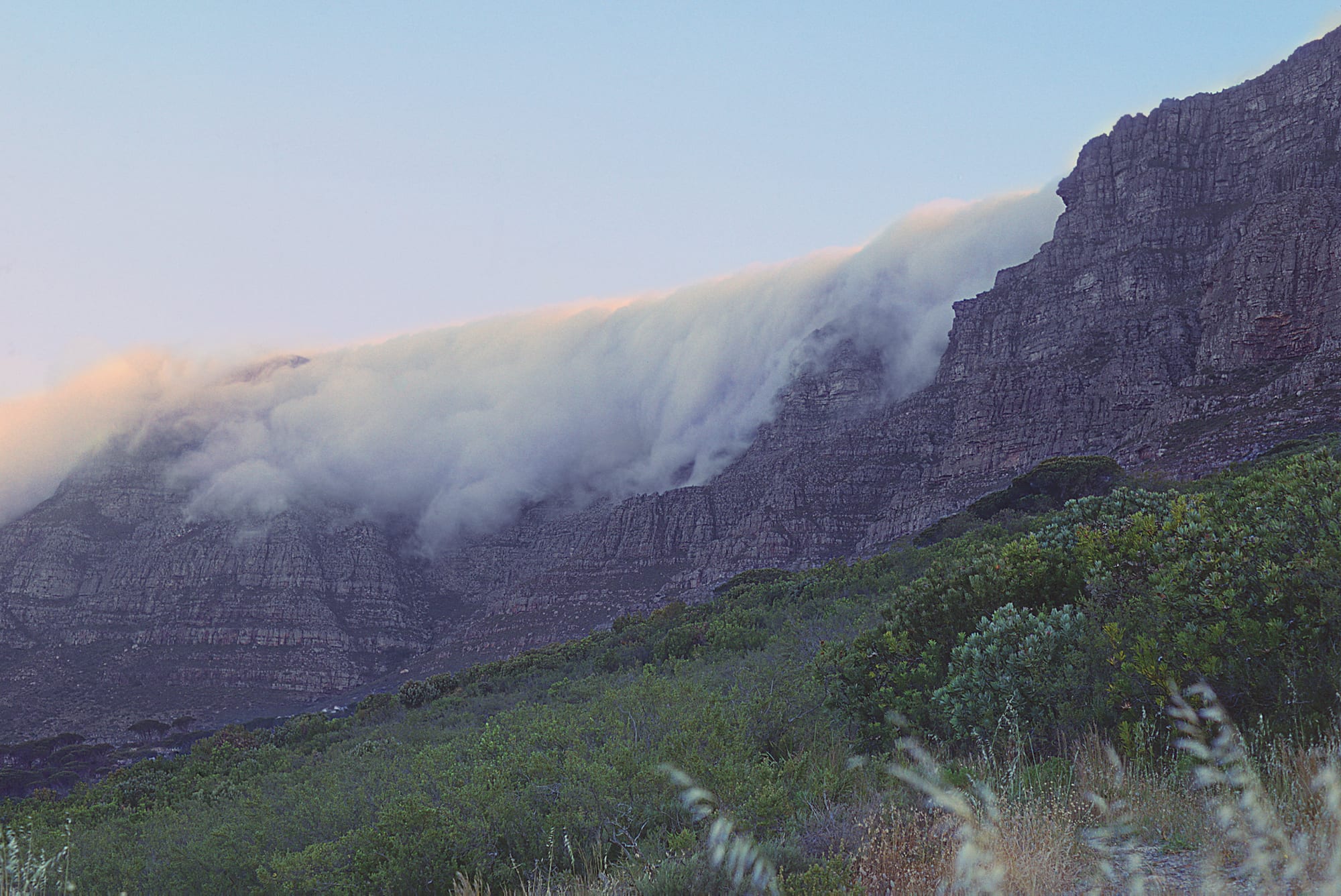 A thick white cloud pouring over the edge of a long line of grey sandstone cliffs and down their face, above slopes of green bush