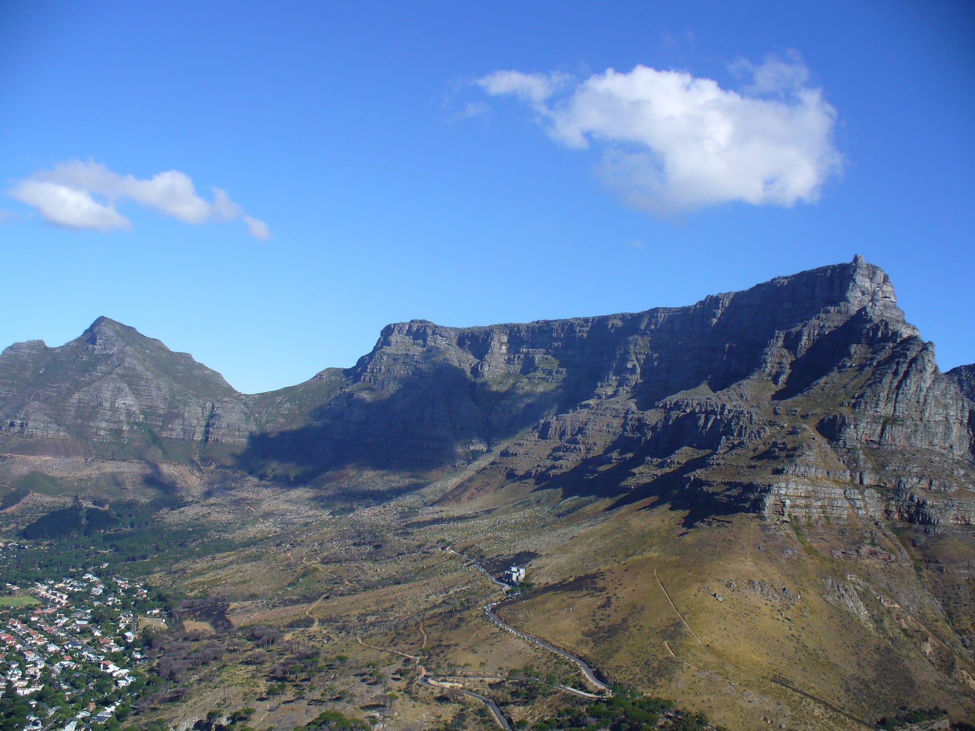 A long flat-topped mountain of grey cliffs under a clear blue sky, with a pointed peak to its left and houses on the lower slopes