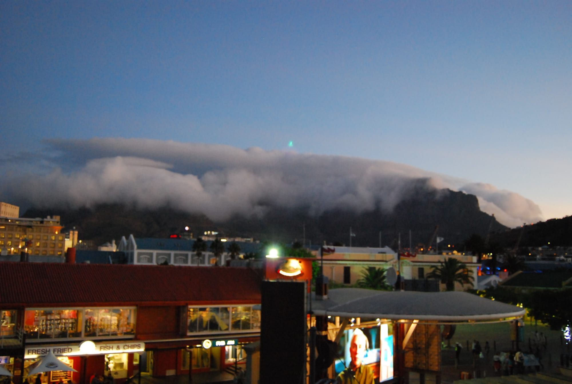 A long rolling cloud covering the top of a dark mountain at dusk, above lit-up shops and restaurants of a harbour-front complex