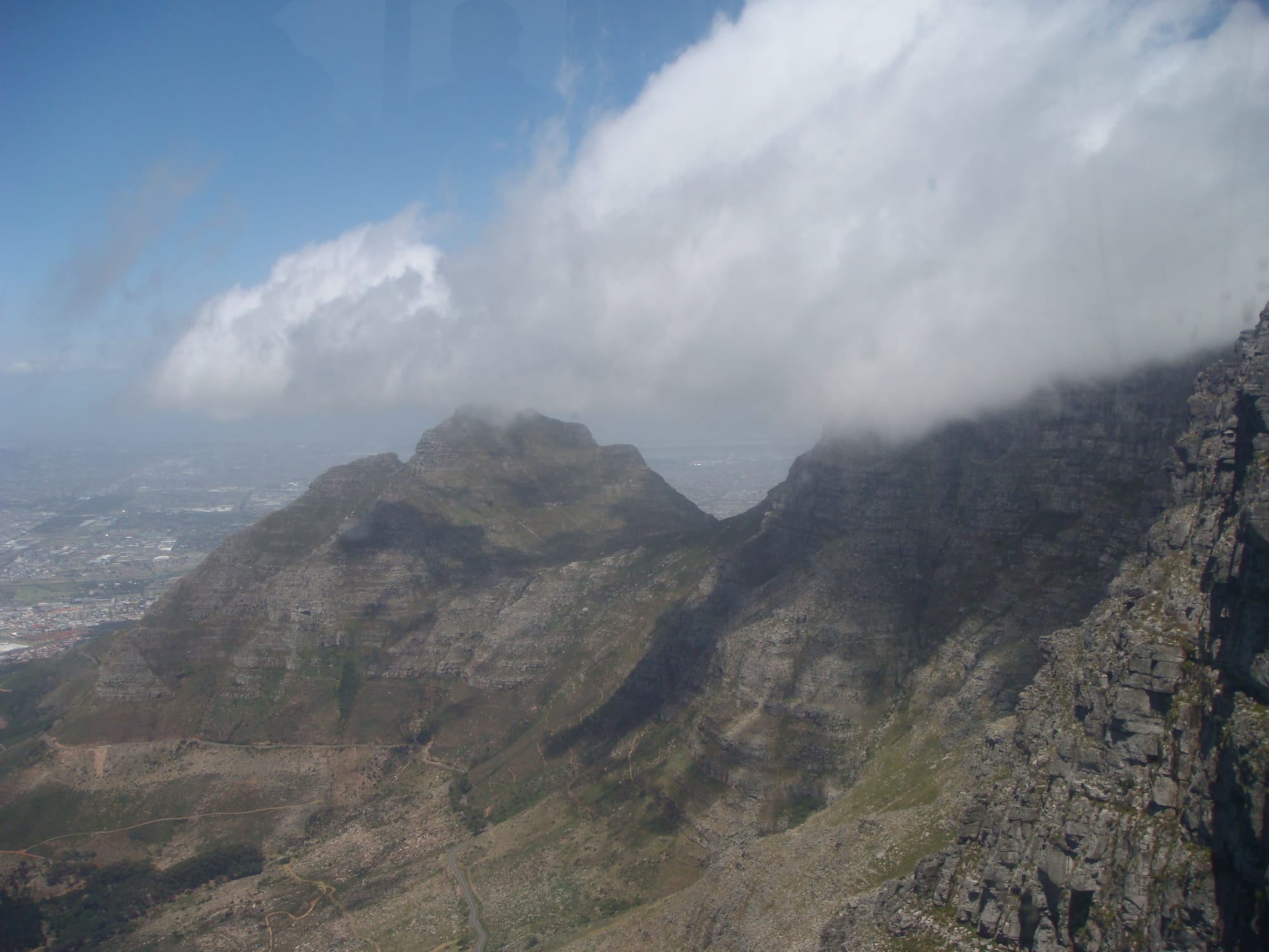 A bank of white cloud lying on a mountain ridge and drifting over a rocky peak, with the city spread out on the plain far below