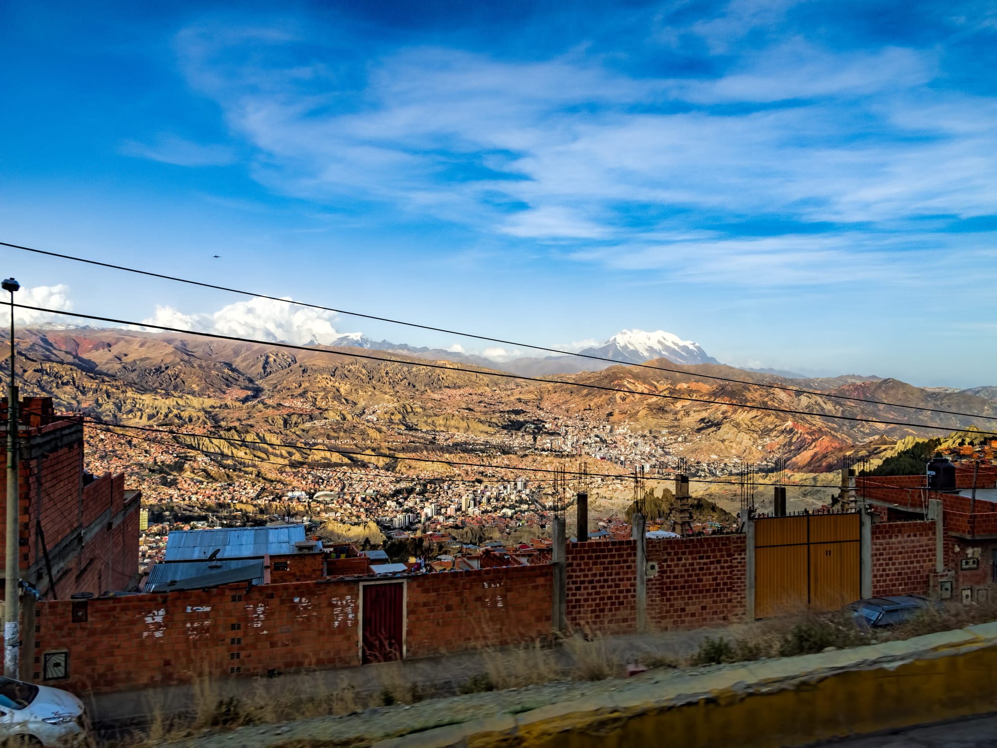 A city of red-brick and white buildings filling a wide, eroded valley below brown hills, with a snow-capped mountain on the horizon under a blue sky