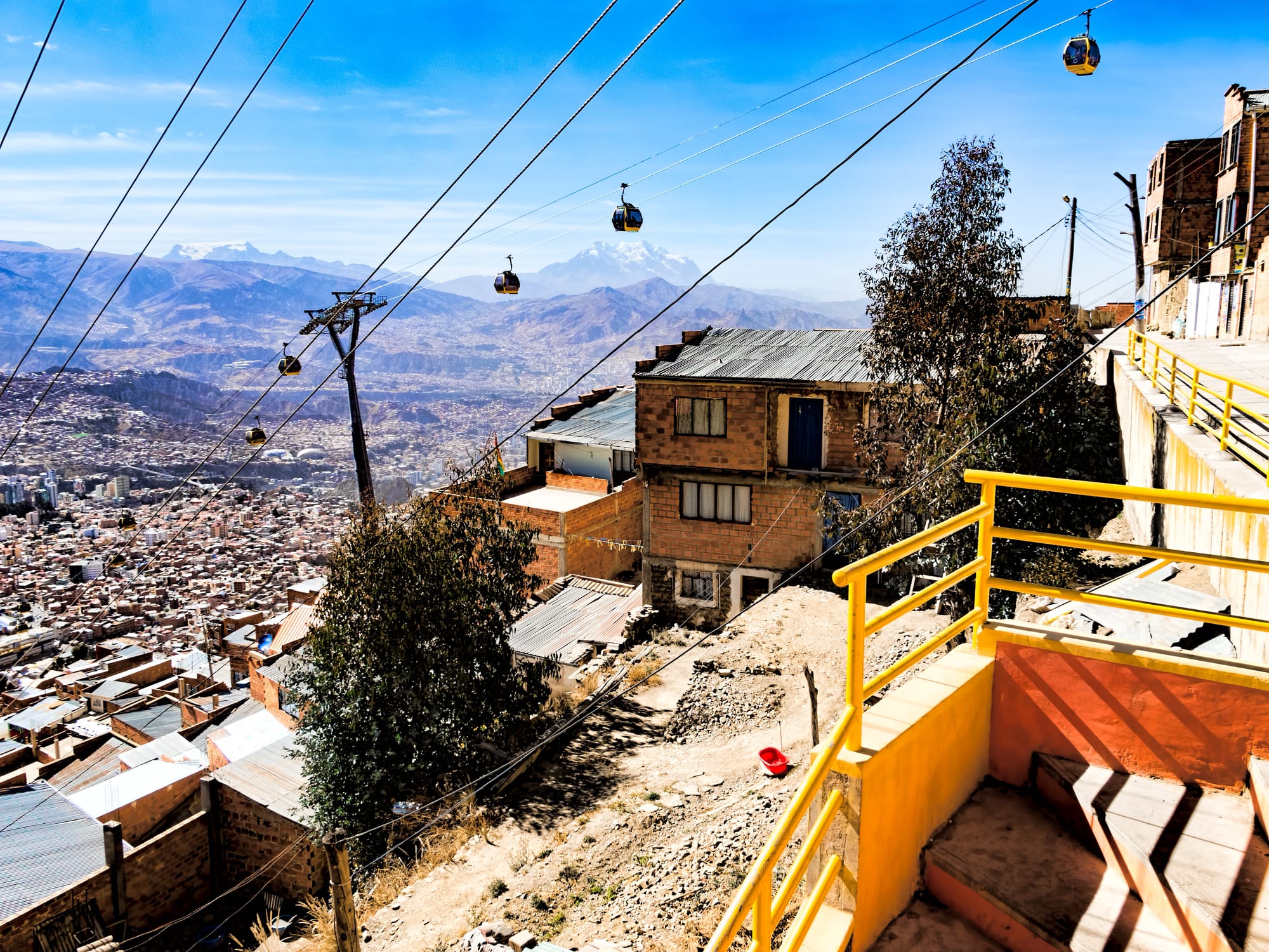 Small yellow cable cars on wires running down from a hillside of brick houses towards a vast city in a valley, with snowy mountains beyond