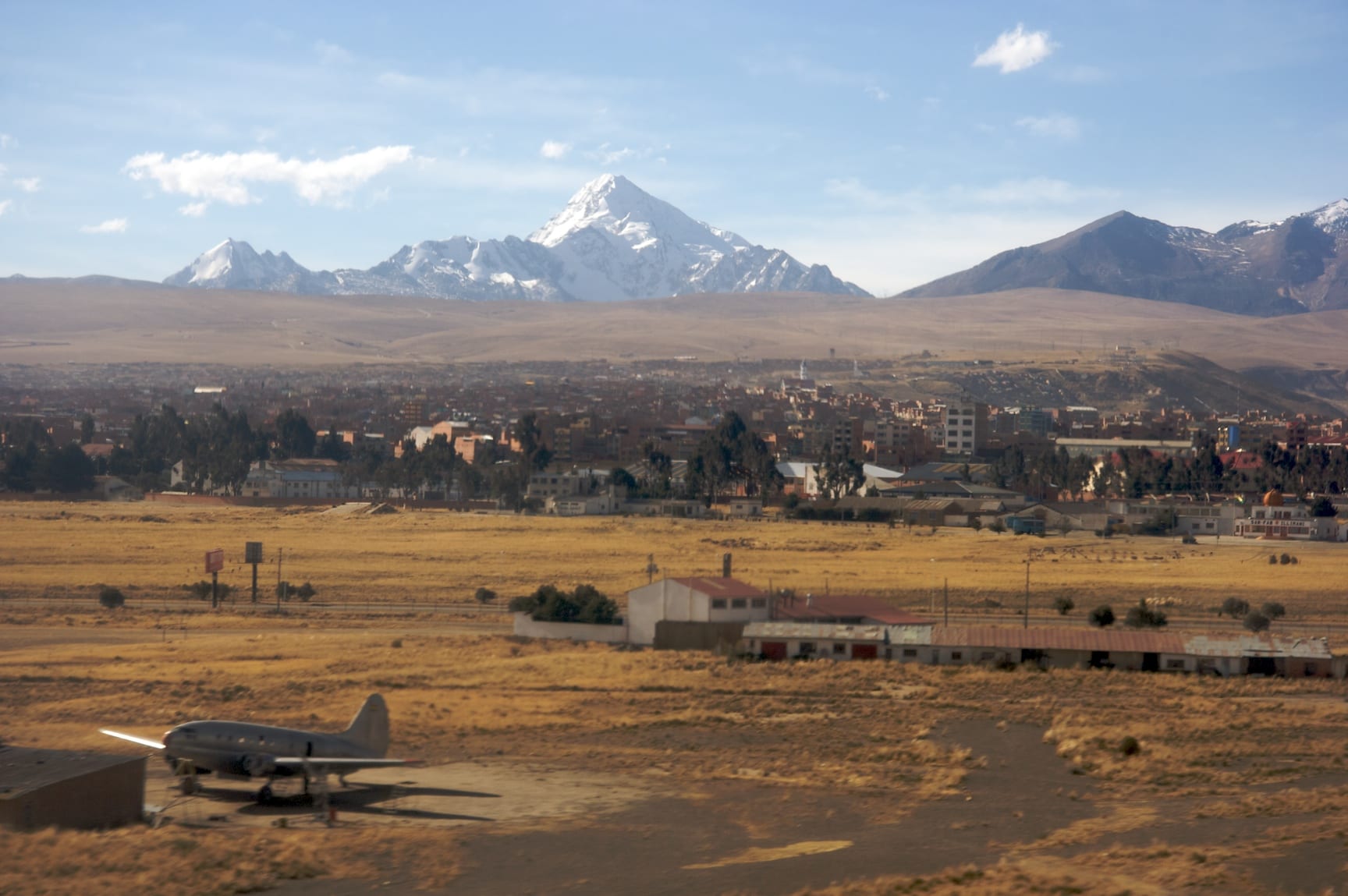 A flat, golden grassland with an old propeller aircraft and low buildings in front of a town, below a snow-covered pyramid-shaped peak