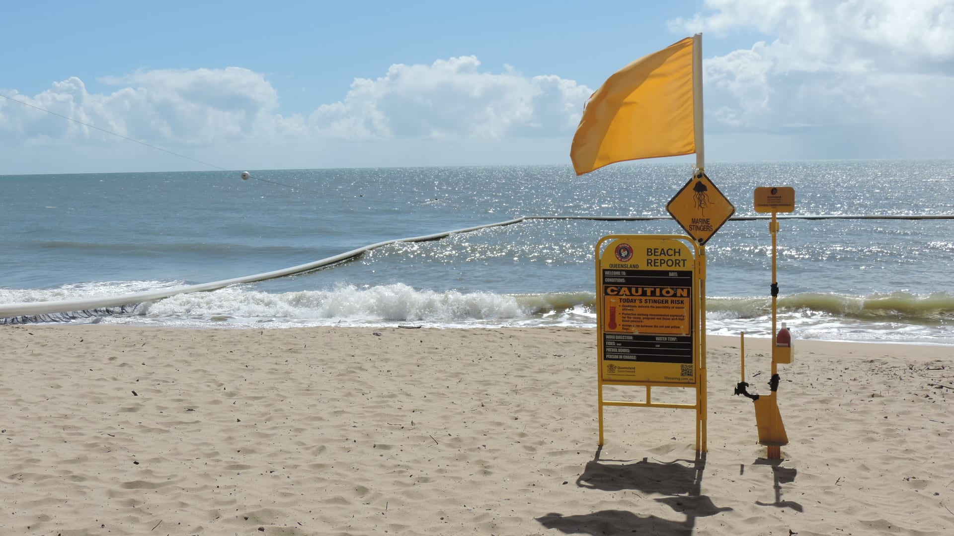 A floating net enclosure in the surf off a sandy beach, with a yellow flag, a stinger warning sign and a post holding a vinegar bottle in the foreground