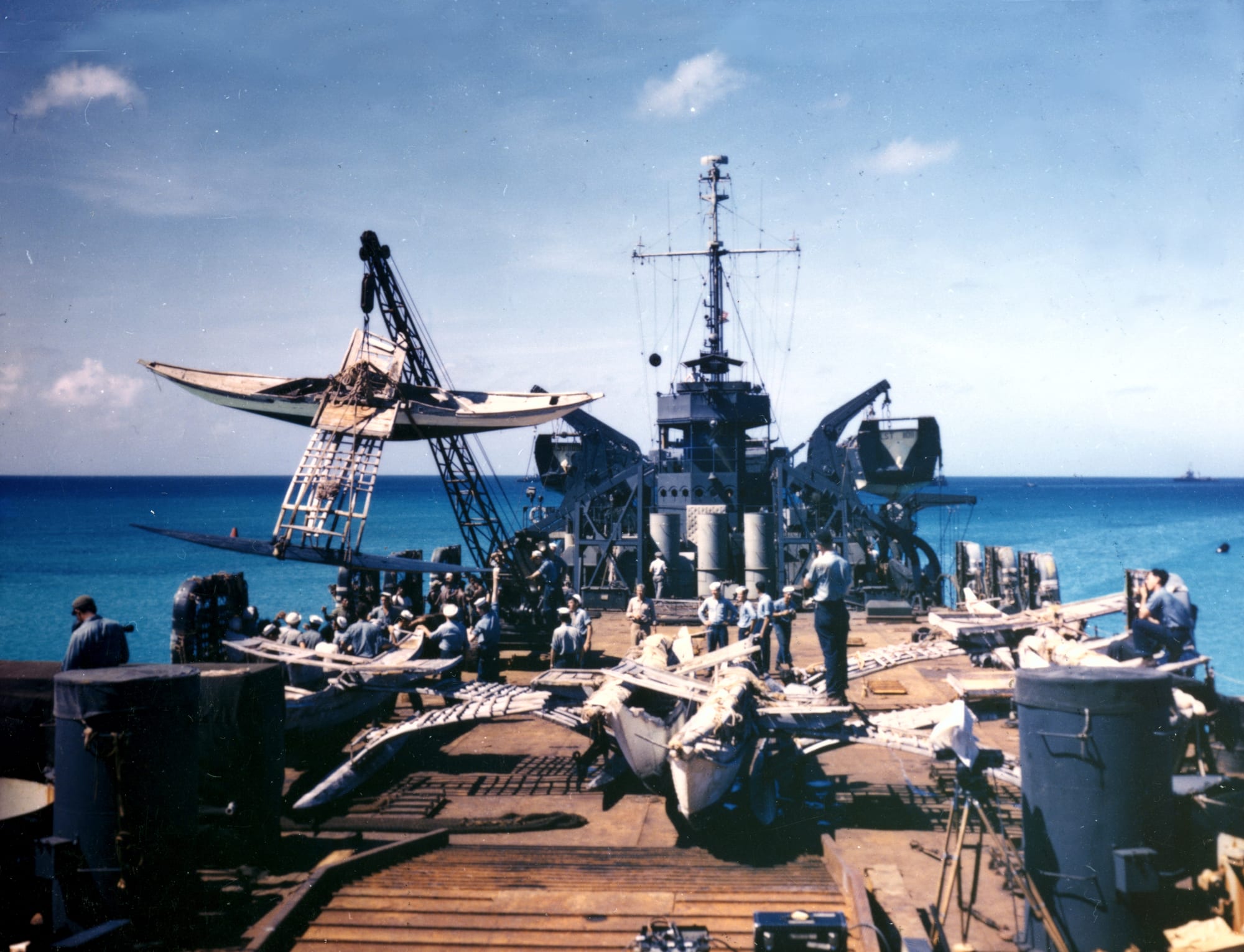 A traditional outrigger canoe being swung by a crane onto the crowded deck of a navy ship, with sailors and more canoes below