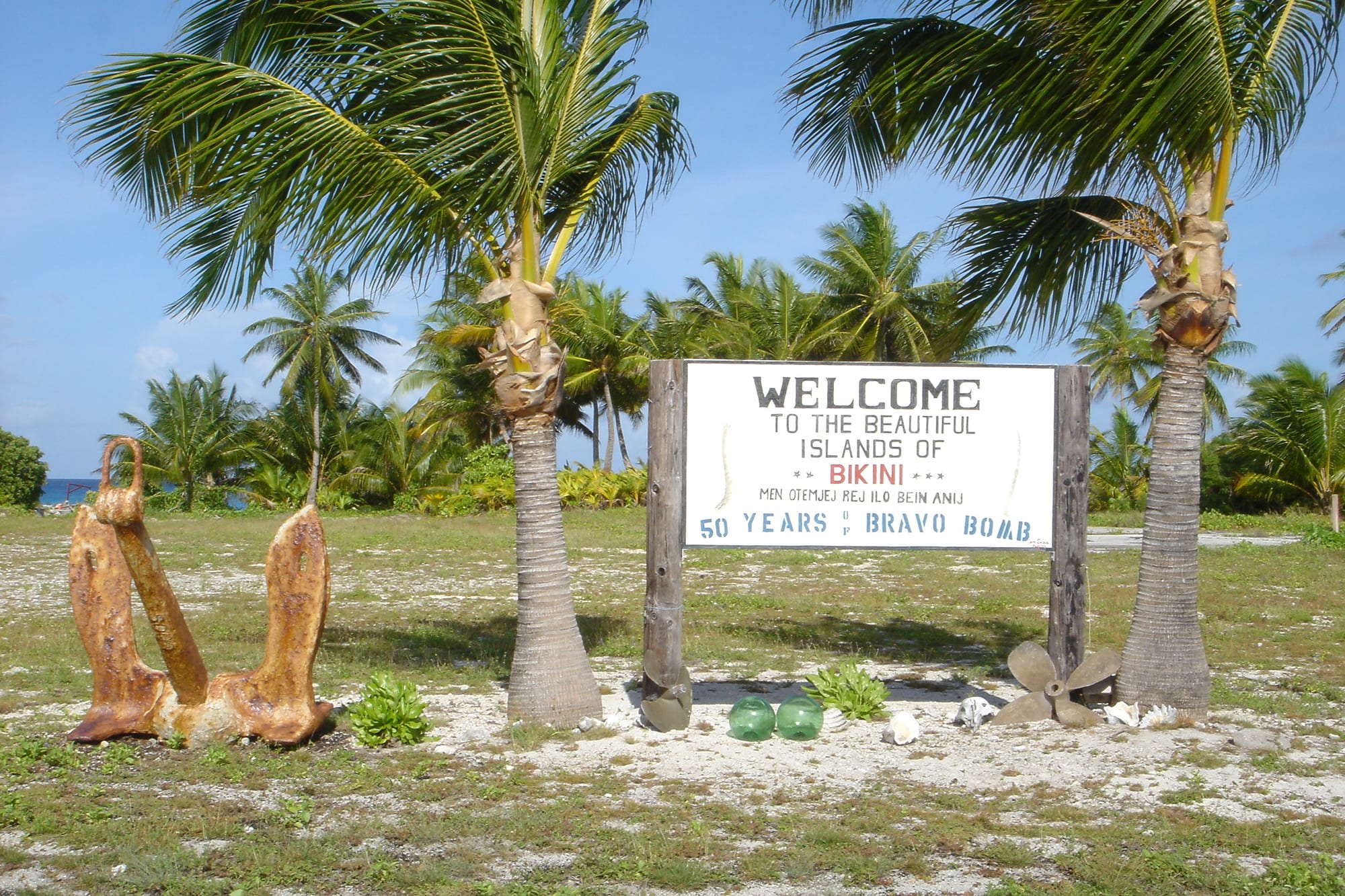 A white wooden sign between two palm trees reading Welcome to the beautiful islands of Bikini, with a rusted anchor and an old propeller beside it