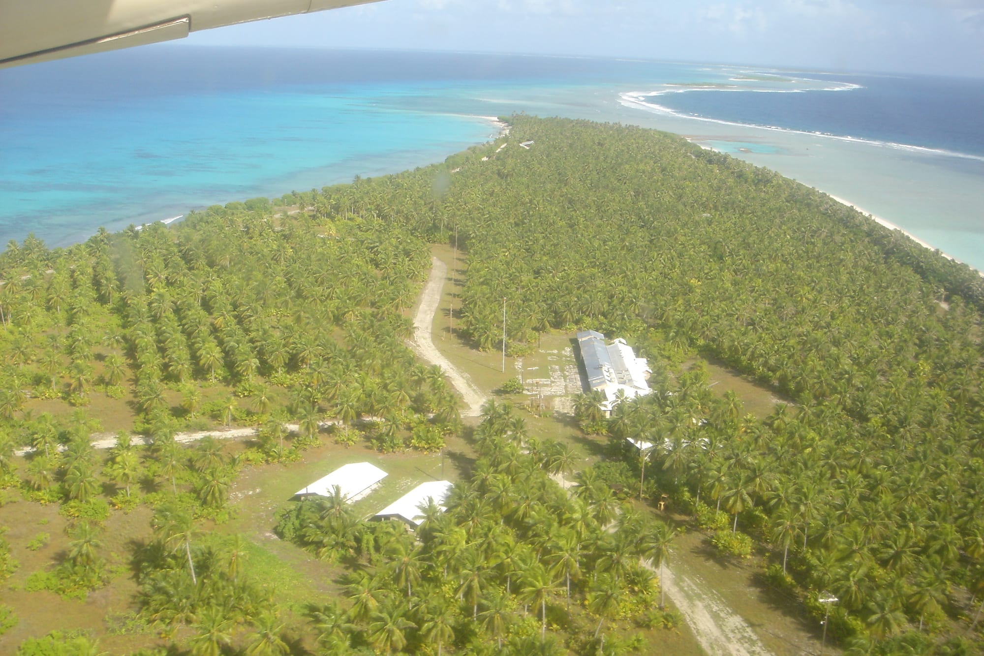 An aerial view of a long island covered in neat rows of coconut palms, with a few white-roofed buildings and a dirt track, between a turquoise lagoon and the open sea