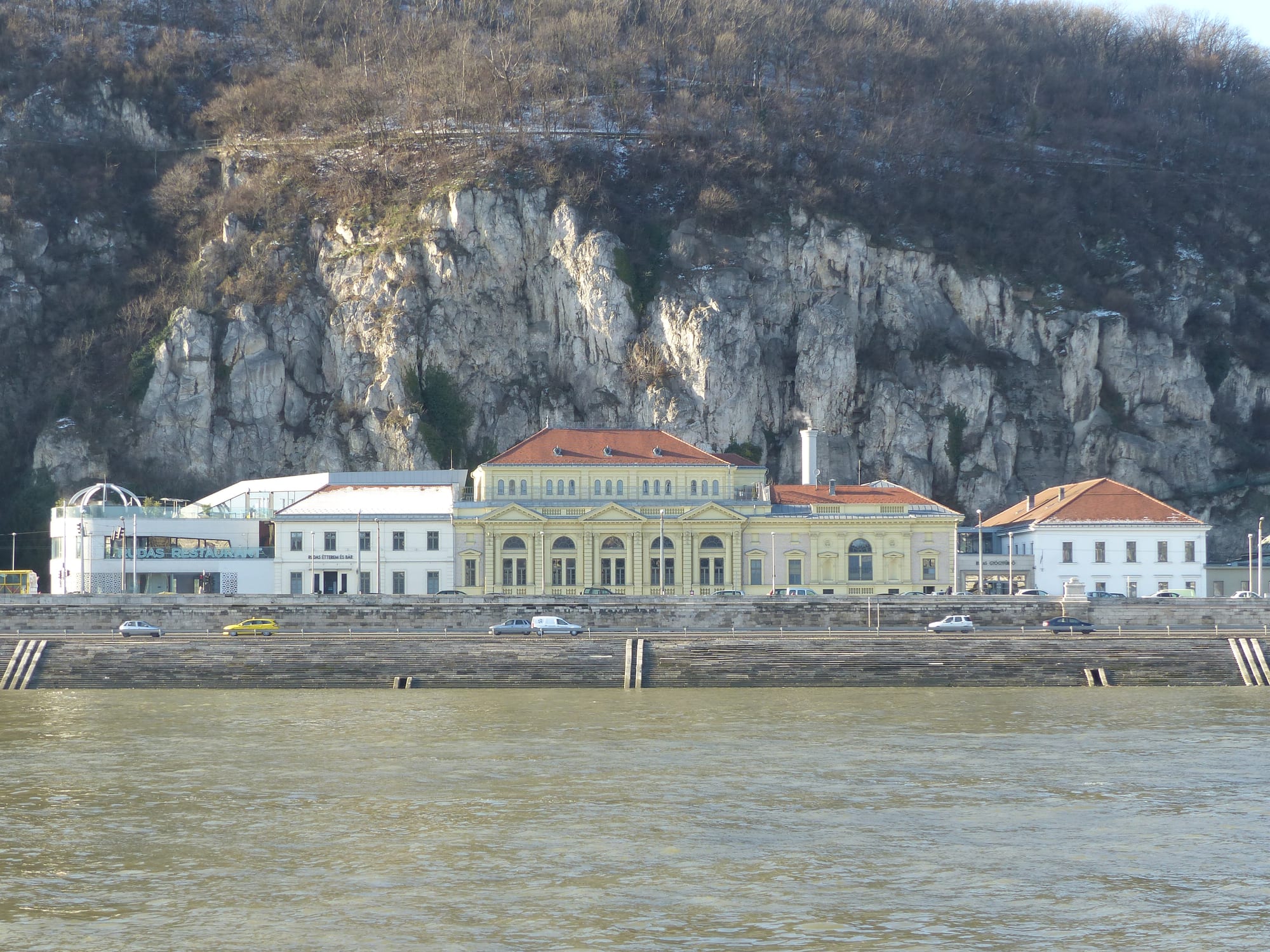A yellow classical building with a red roof on a river embankment, at the foot of a steep wall of pale limestone cliffs