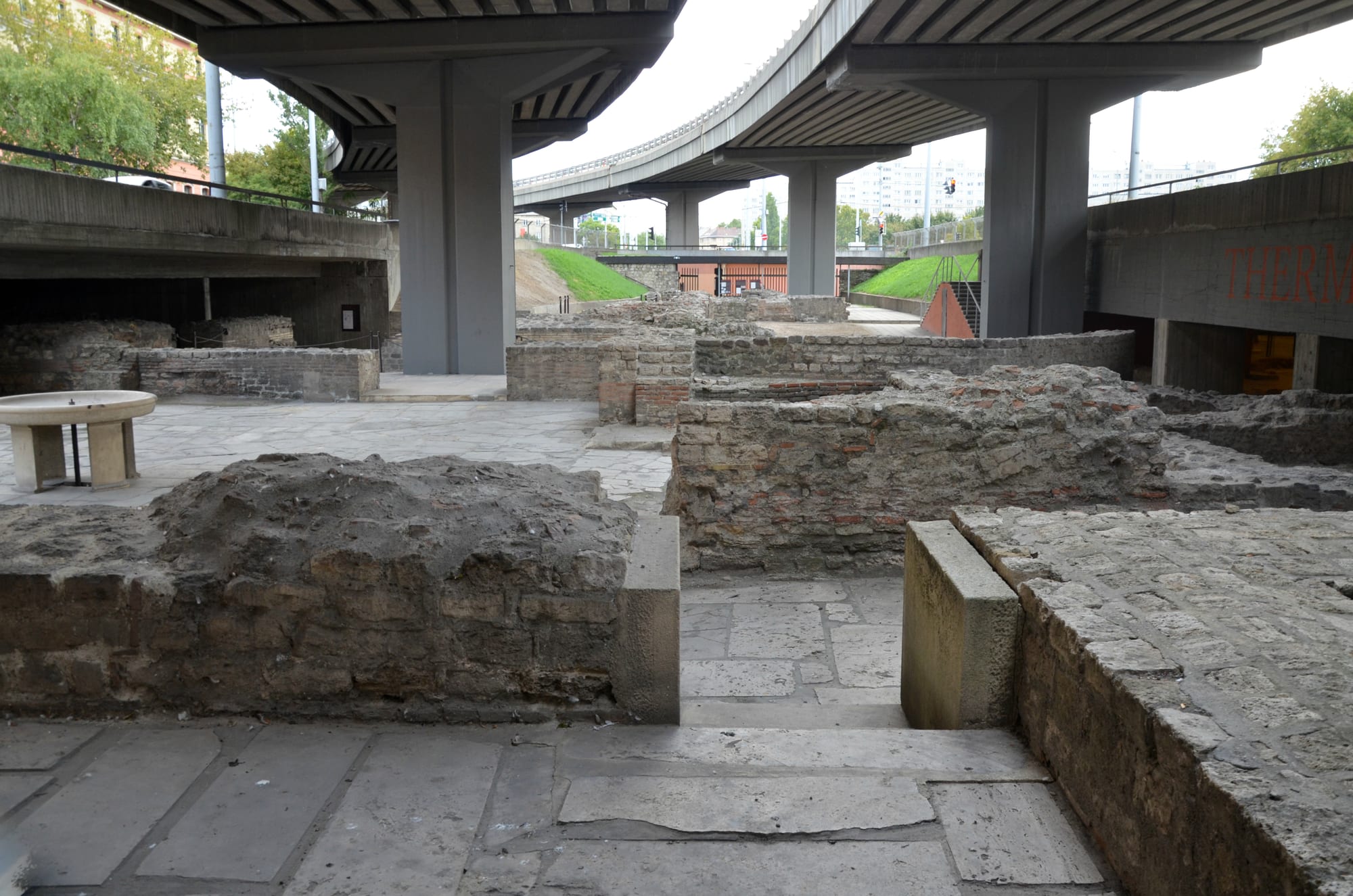 Low brick and stone walls of an excavated Roman building, with paved floors, beneath the concrete pillars of a road flyover