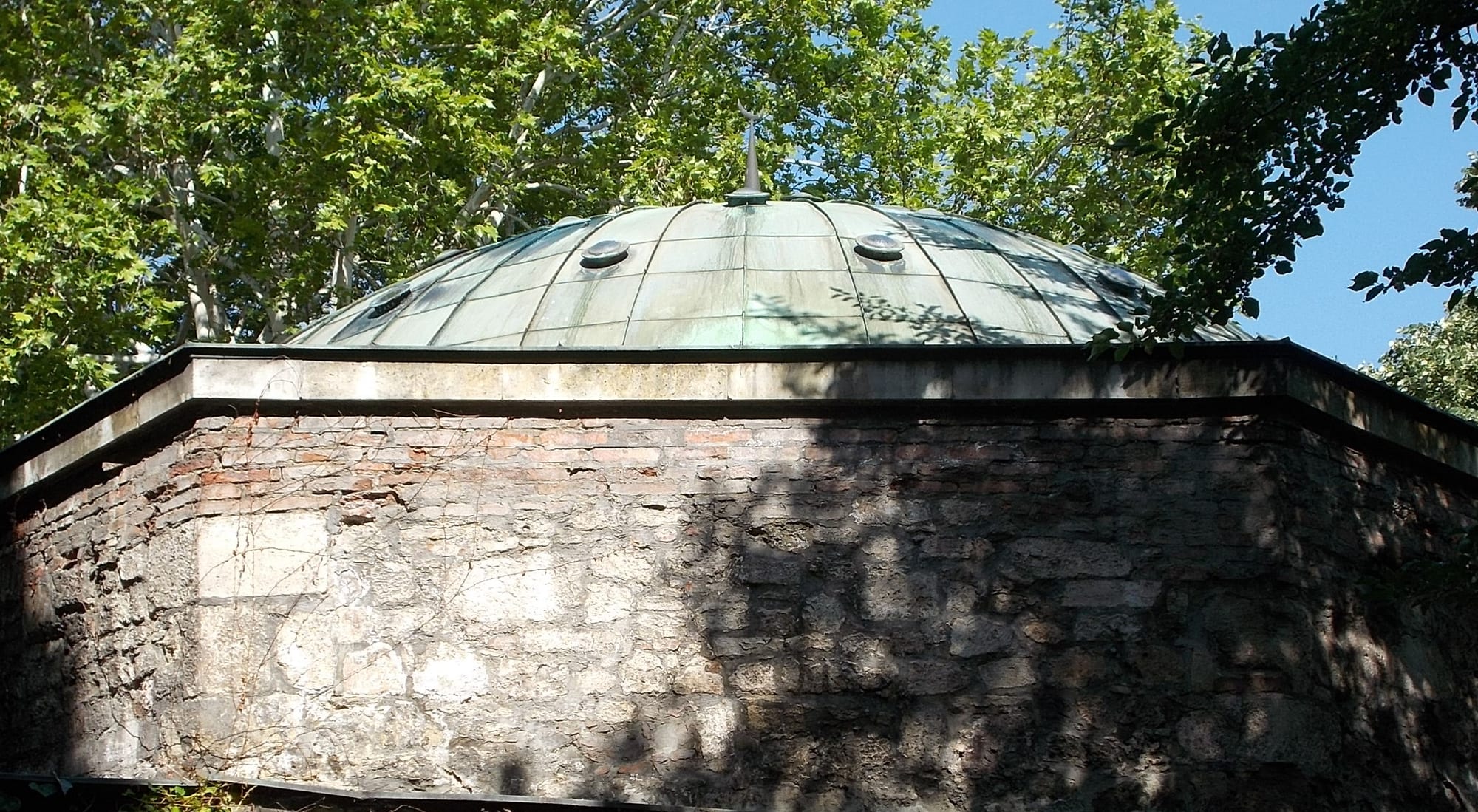 A low copper-green dome dotted with small round skylights, on top of an octagonal stone and brick wall, among trees