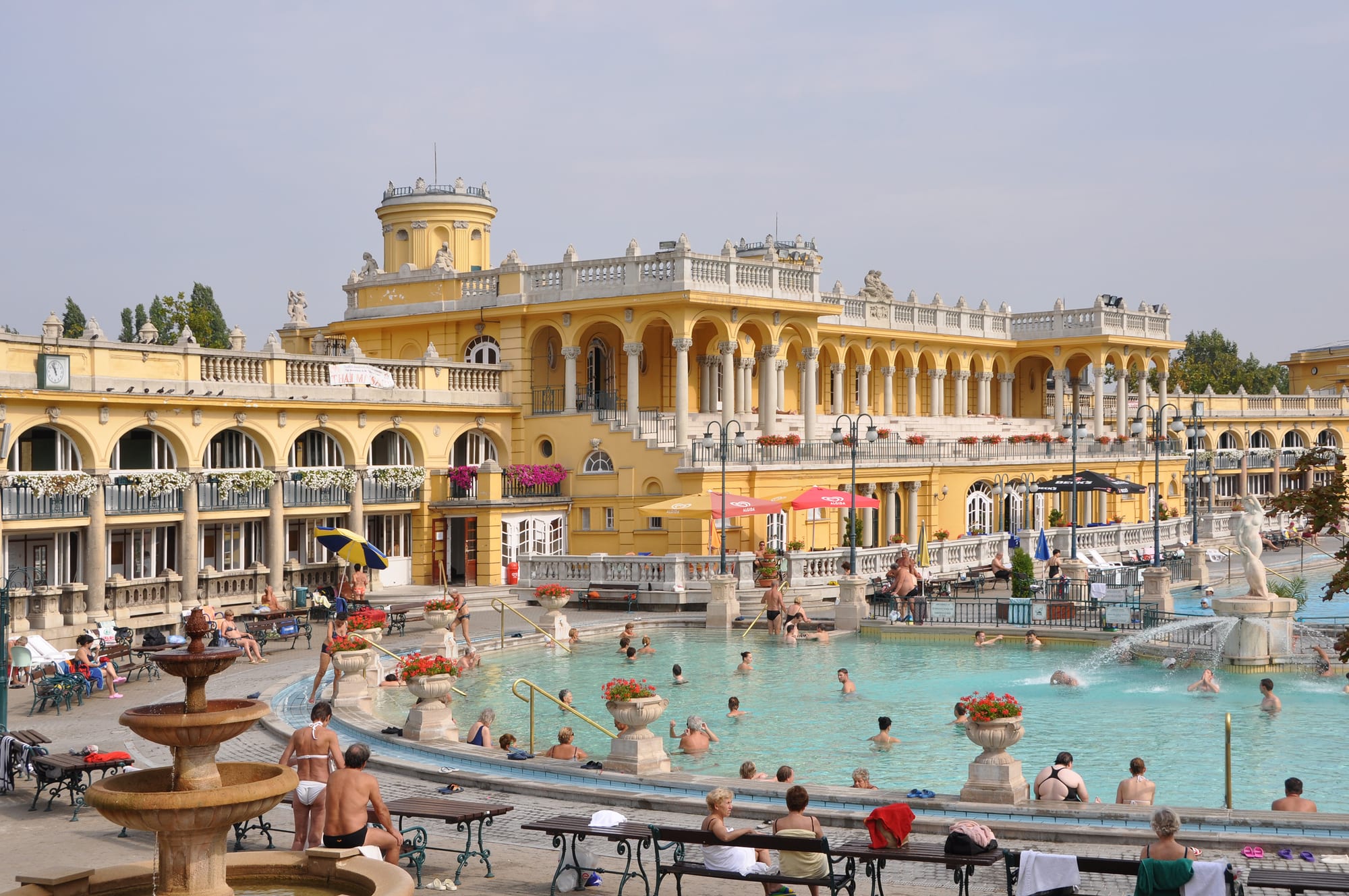 Bathers in a large outdoor pool of turquoise water surrounded by a grand yellow neo-baroque building with arcades, balustrades and statues