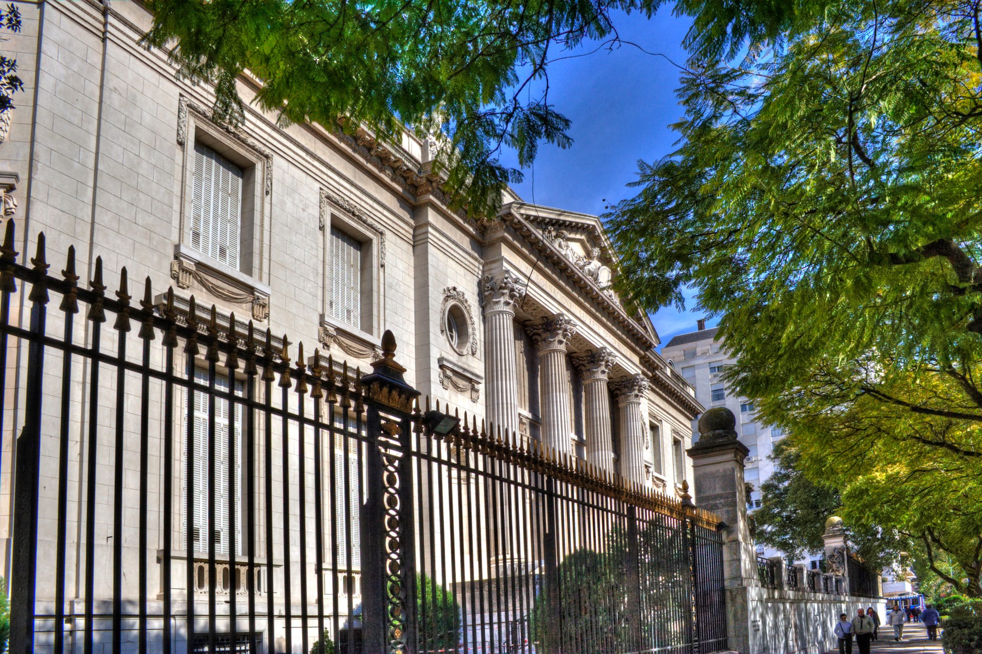 The pale stone side of a neoclassical mansion with tall Corinthian columns and shuttered windows, behind black iron railings and green trees