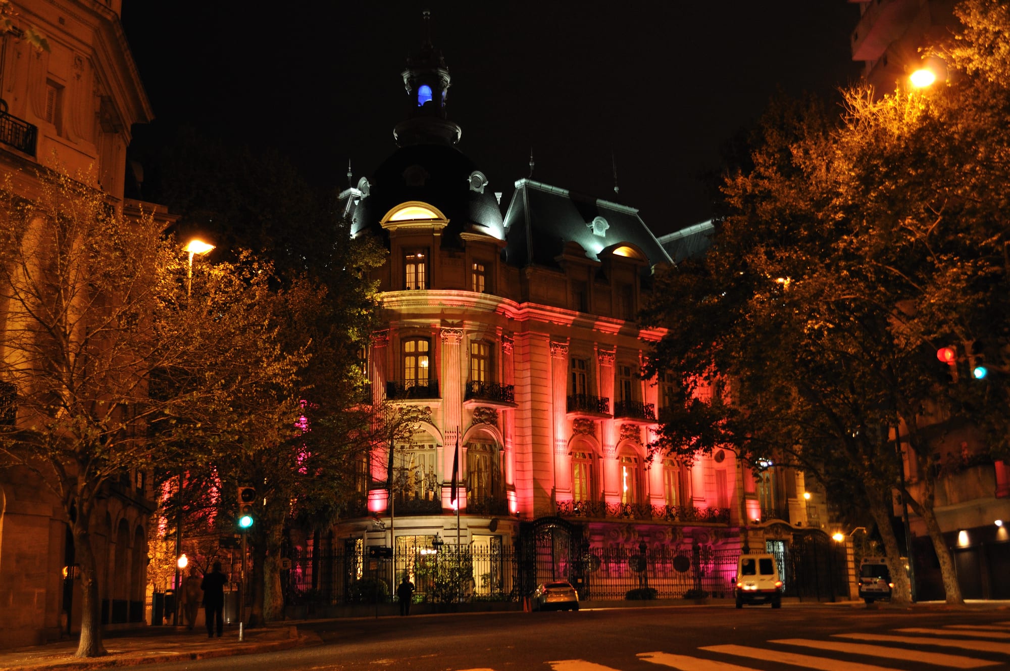 An ornate mansion with a domed corner tower and steep slate mansard roof, lit pink and gold at night behind iron railings