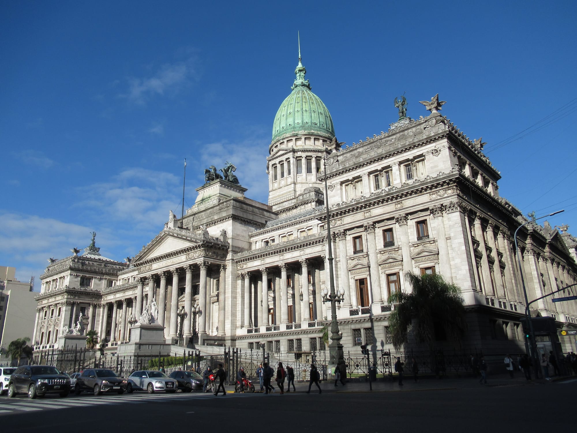 A grand white neoclassical palace with columns, rooftop statues and a tall green copper dome under a blue sky