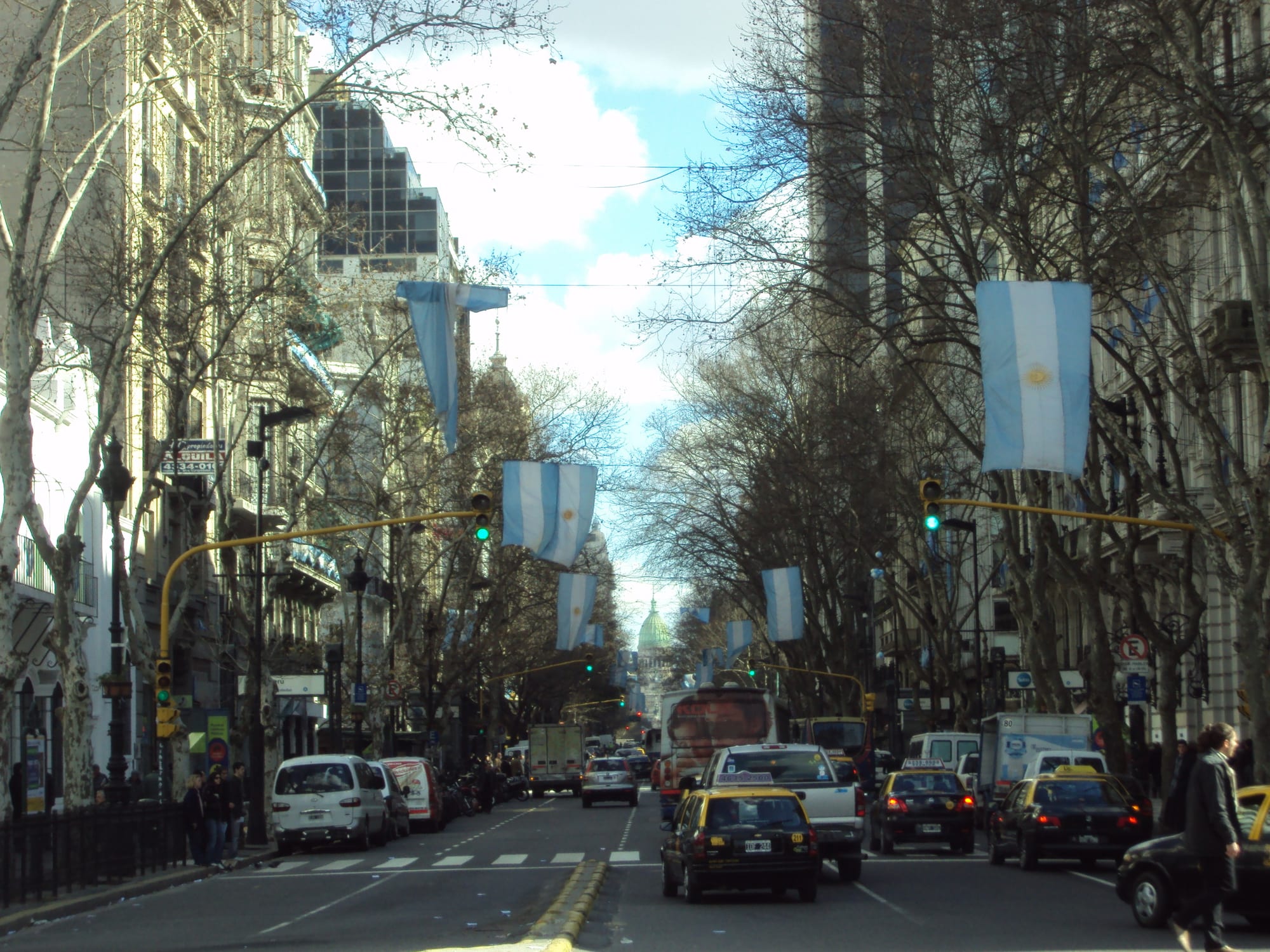 A tree-lined avenue of ornate stone buildings hung with blue and white flags, with yellow-topped taxis and a green dome at the far end