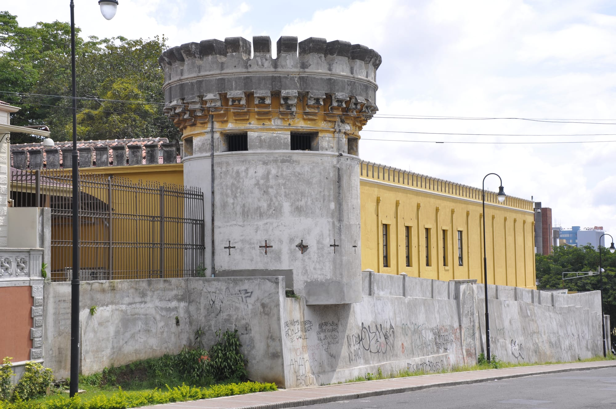 A grey crenellated corner tower and high wall attached to a long yellow building on a city street