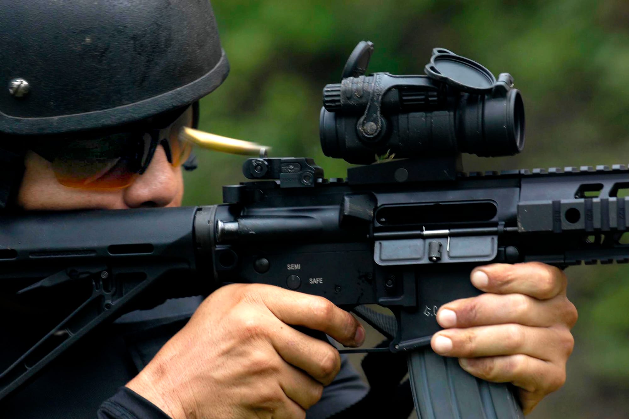 Close view of a helmeted man aiming a black rifle as a spent cartridge flies from it