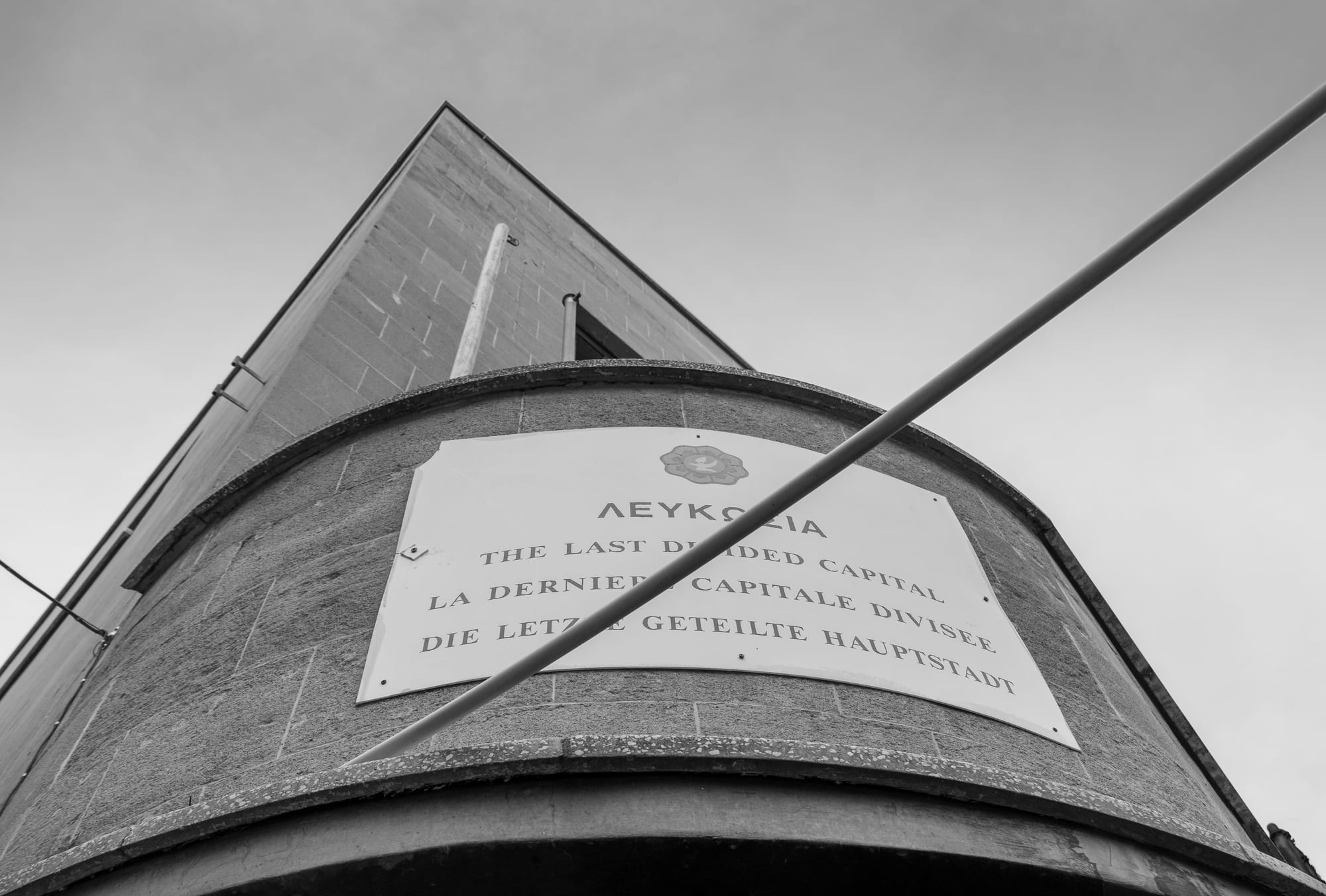 A black and white view looking up at a rounded stone building with a sign reading The Last Divided Capital in Greek, English, French and German