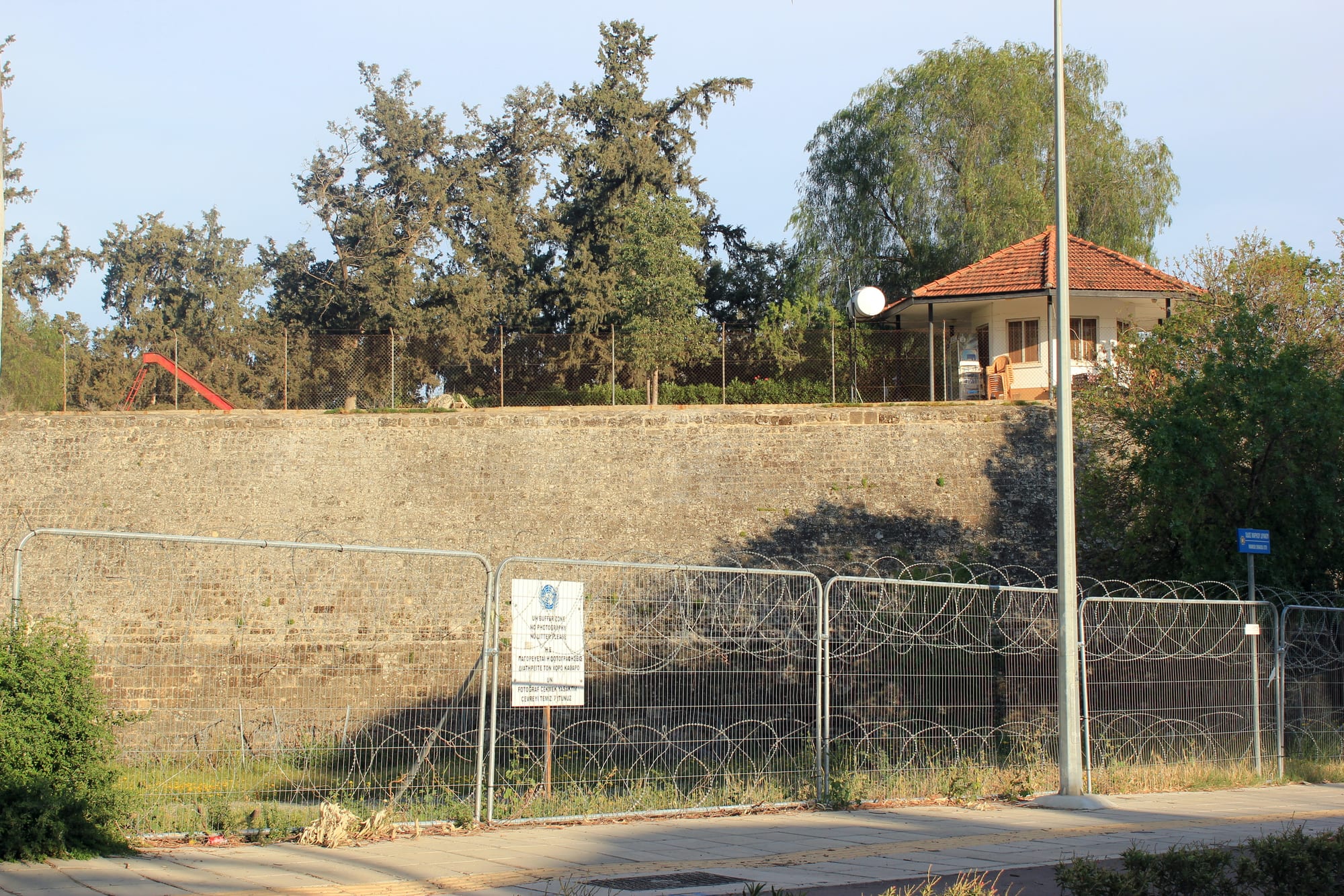 An old stone fortification wall behind a temporary metal fence hung with razor wire and a white United Nations sign