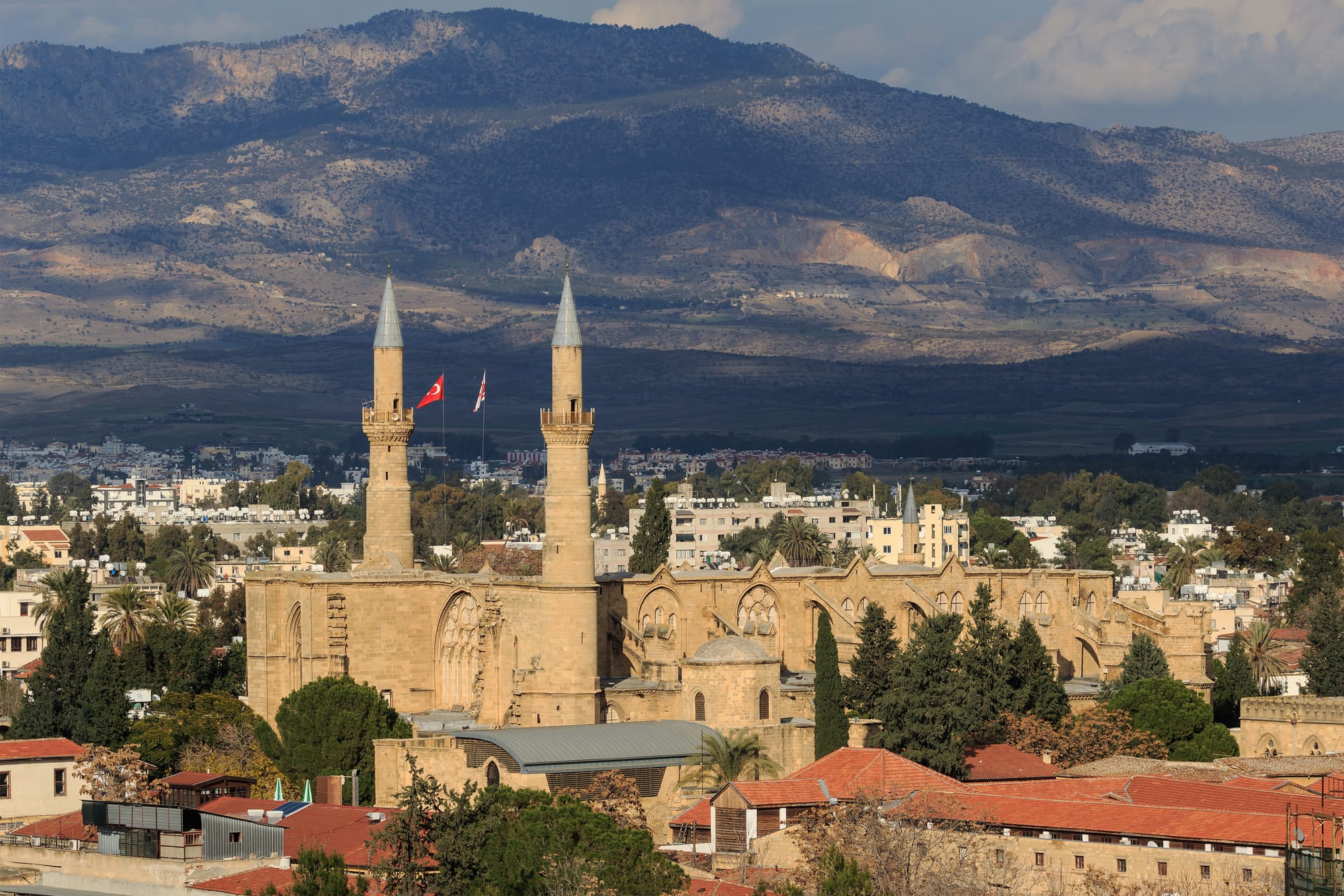 A Gothic cathedral with two tall minarets added to its facade, surrounded by low rooftops, with brown mountains in the distance