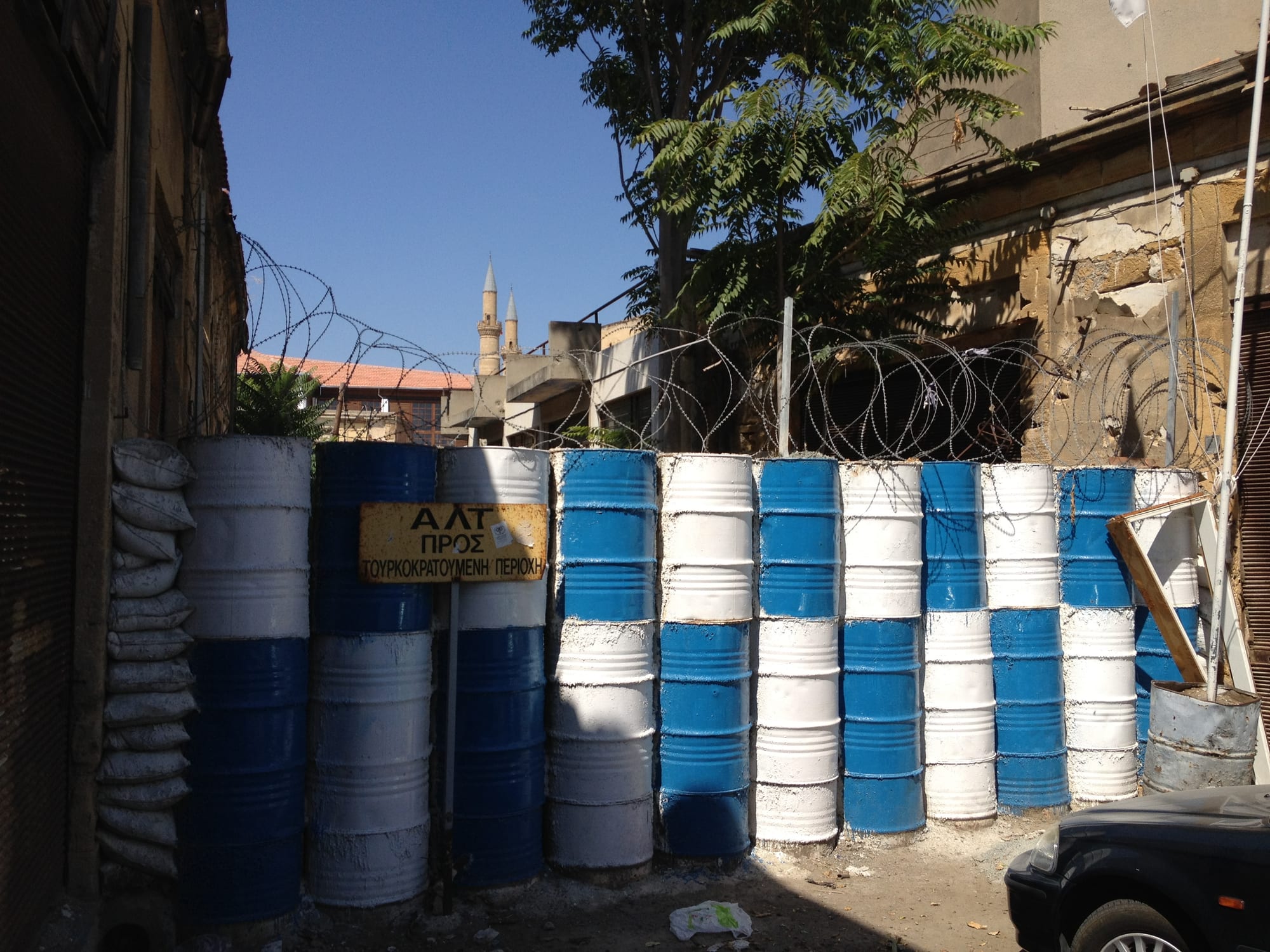 A wall of blue and white painted oil drums topped with coils of barbed wire blocking a narrow street, with a yellow sign in Greek and two minarets rising behind