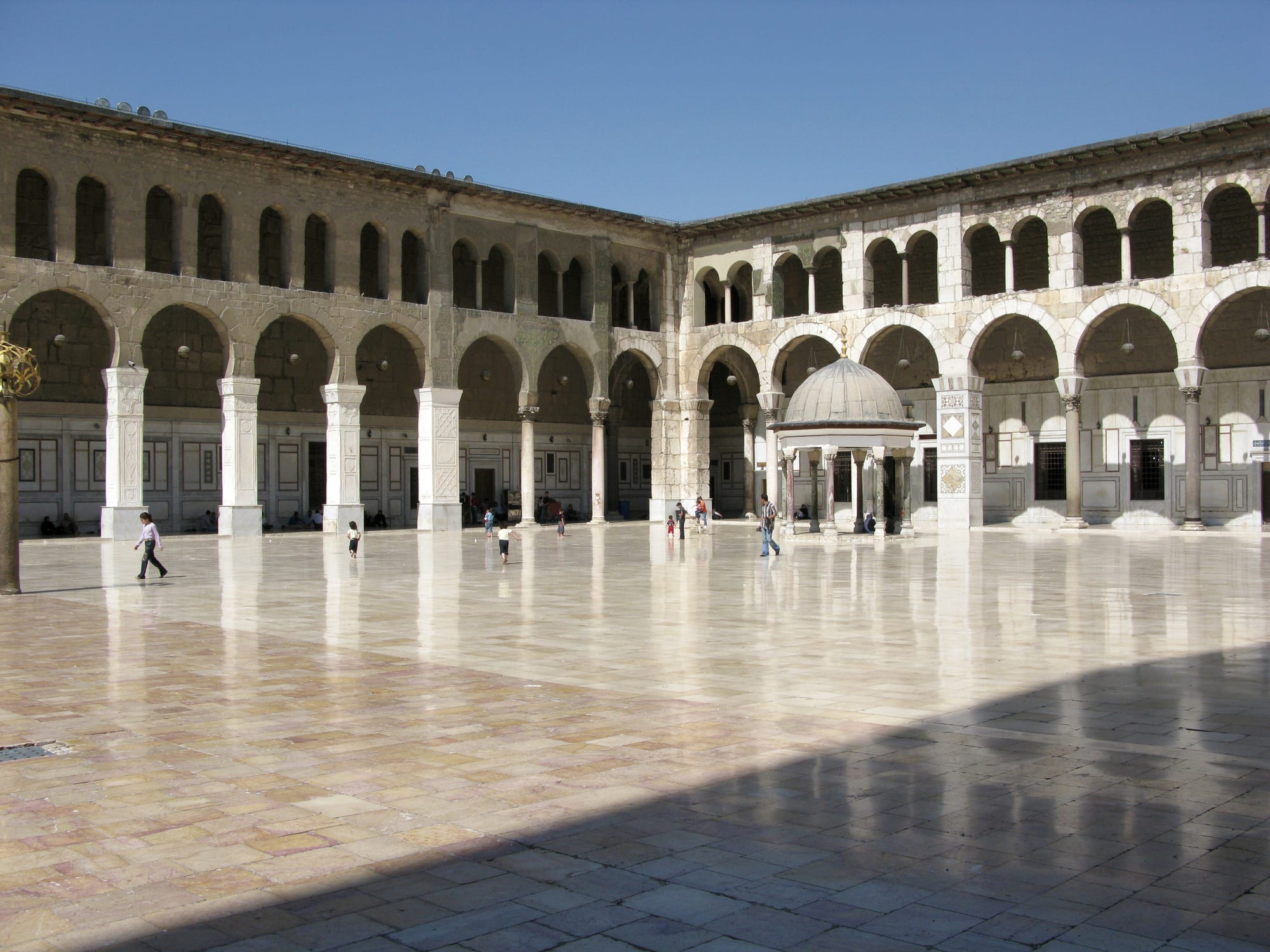 A huge courtyard paved in polished marble, surrounded by two-storey arcades, with a small domed pavilion and people walking across it