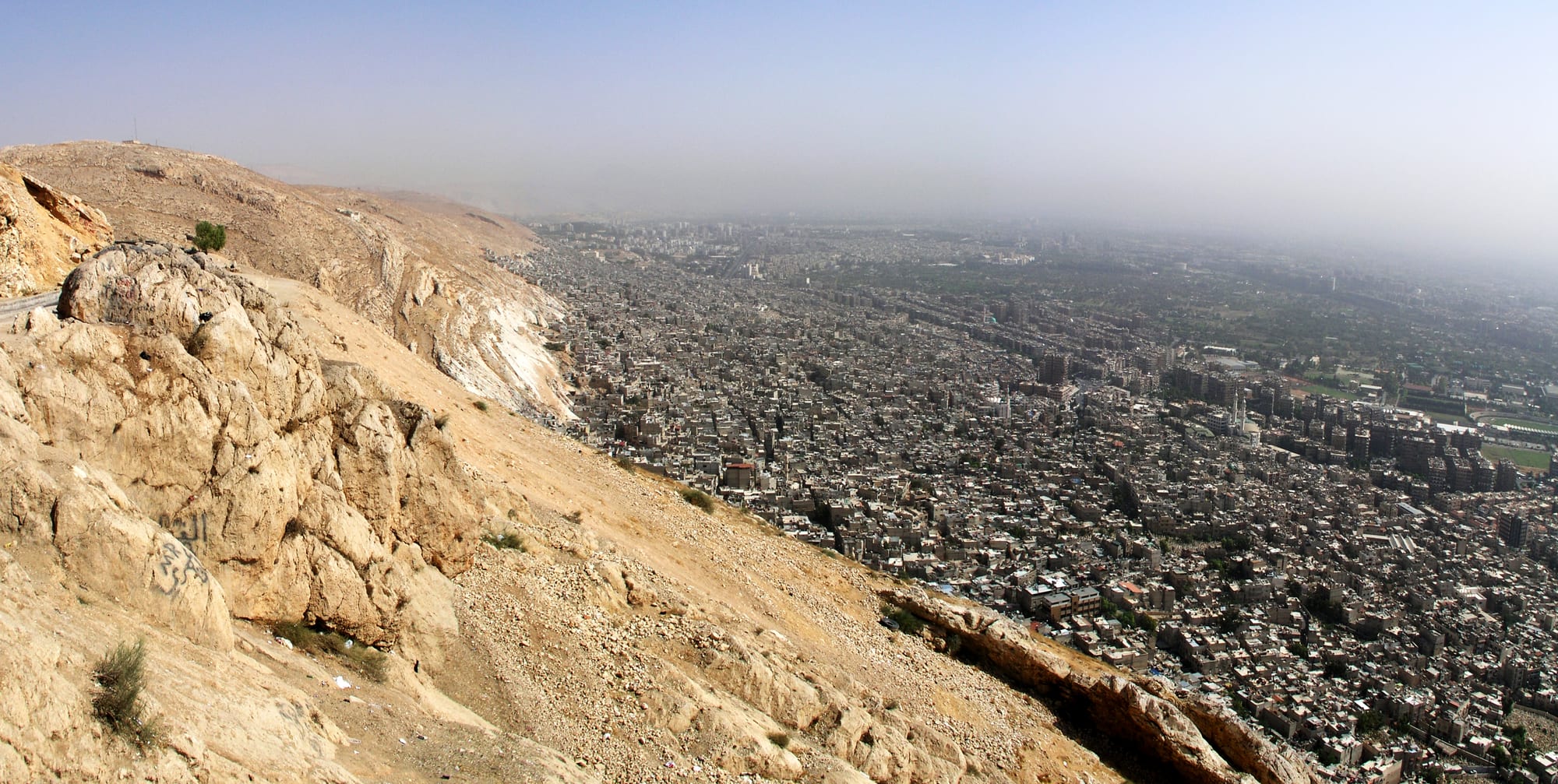 A vast, dense city of low pale buildings spreading across a hazy plain, seen from a bare rocky mountainside