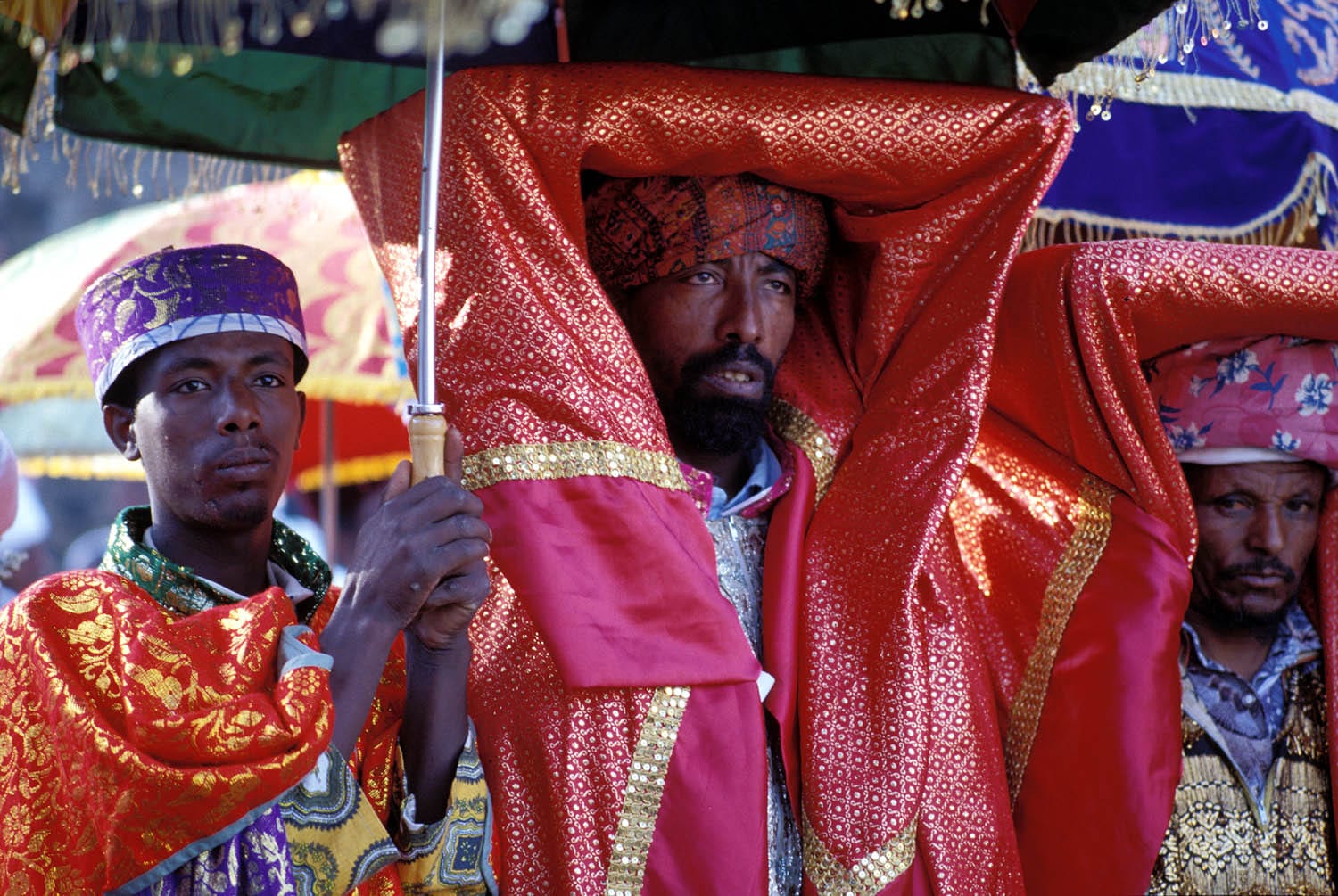 An Ethiopian Orthodox priest carries a sacred object on his head, wrapped in red and gold brocade, beside deacons under embroidered umbrellas