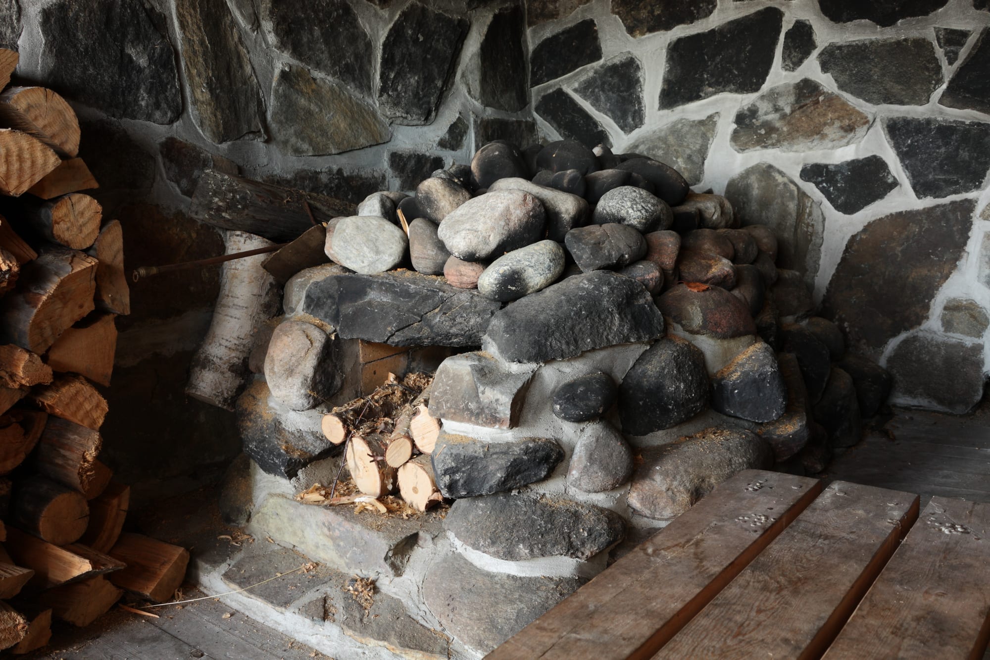 A large heap of rounded grey stones stacked over a firebox filled with split birch logs, beside wooden benches inside a stone-walled room