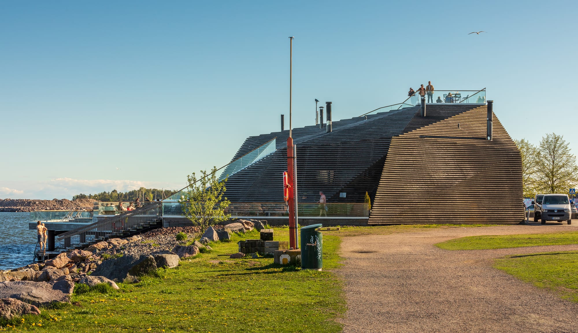 A modern angular building clad in horizontal wooden slats on a rocky seashore, with people on its rooftop terrace and a bather on steps leading into the sea