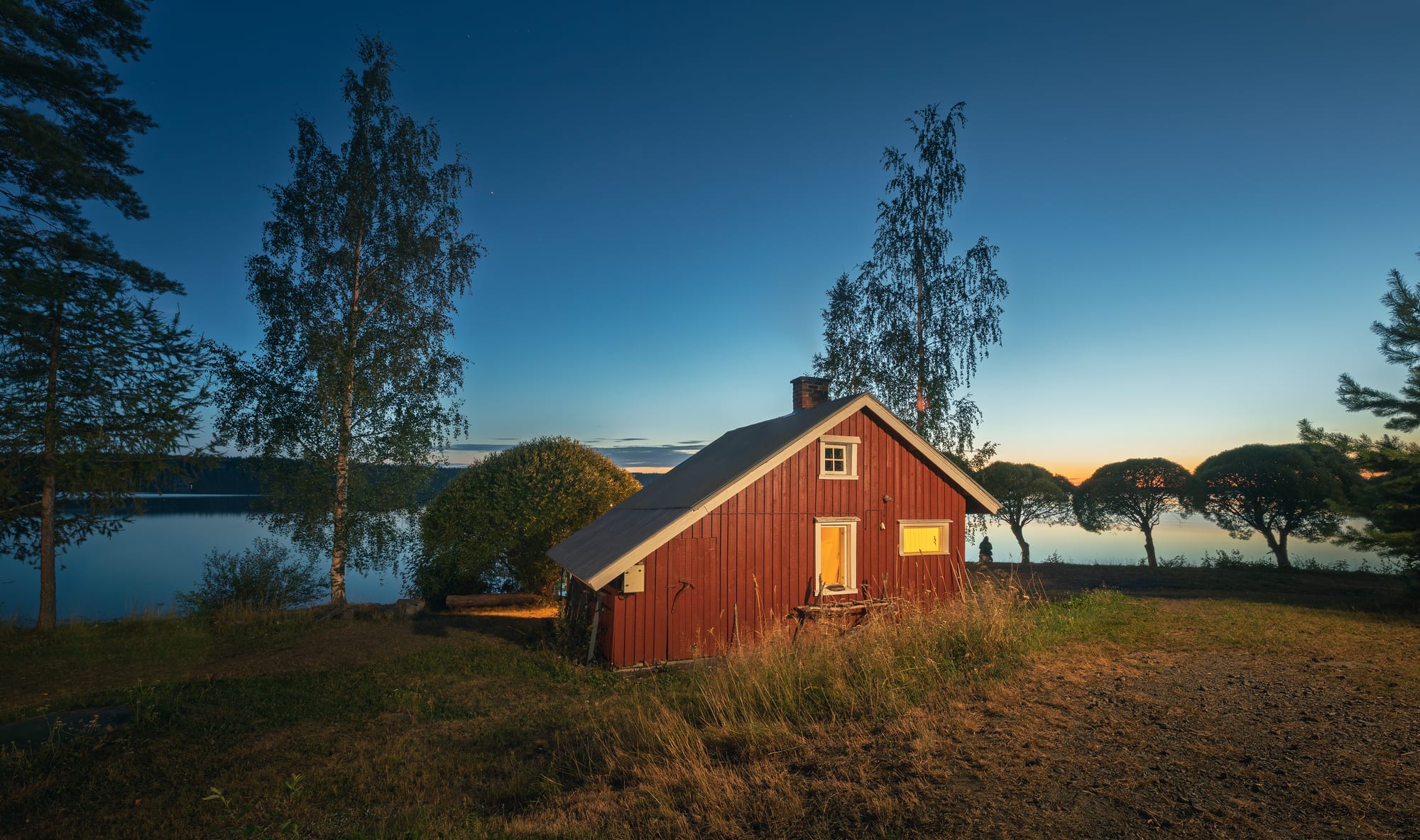 A small red wooden sauna cabin with lit windows on a grassy shore, framed by birch and pine trees, with a calm lake glowing under a twilight sky behind