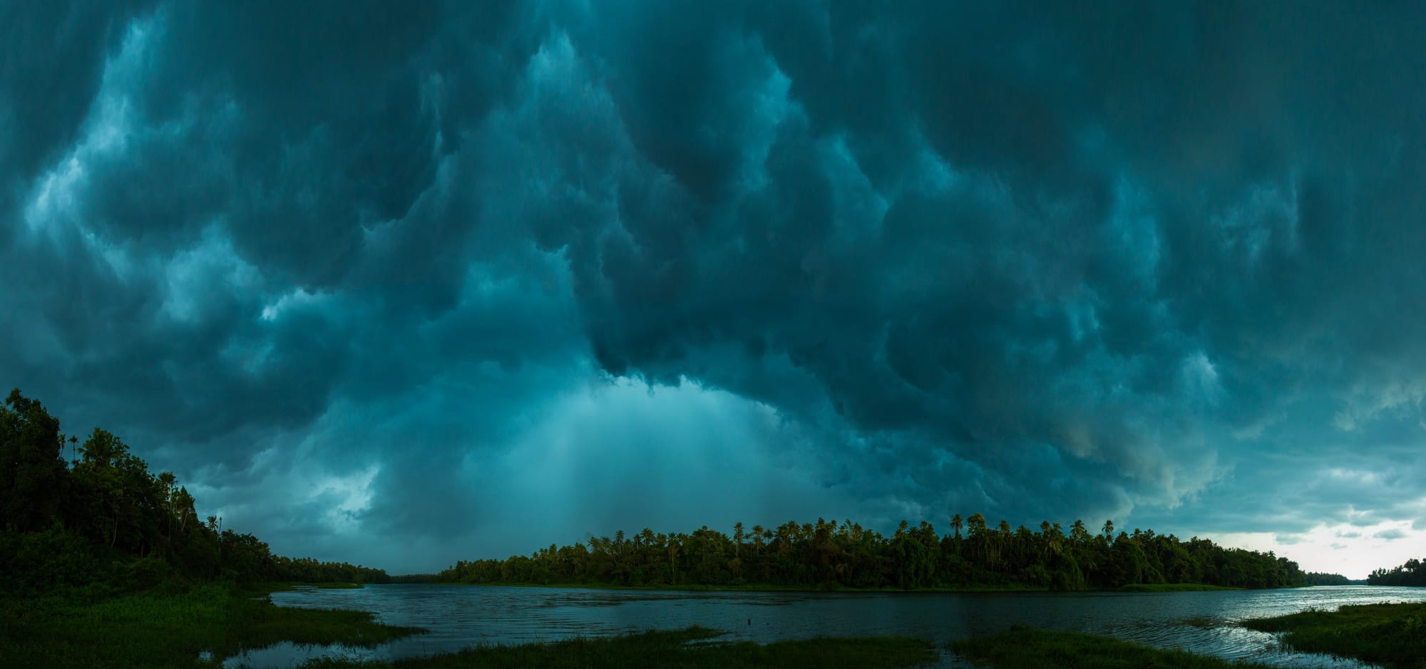 Dark, heavy storm clouds filling the sky above a calm river lined with coconut palms