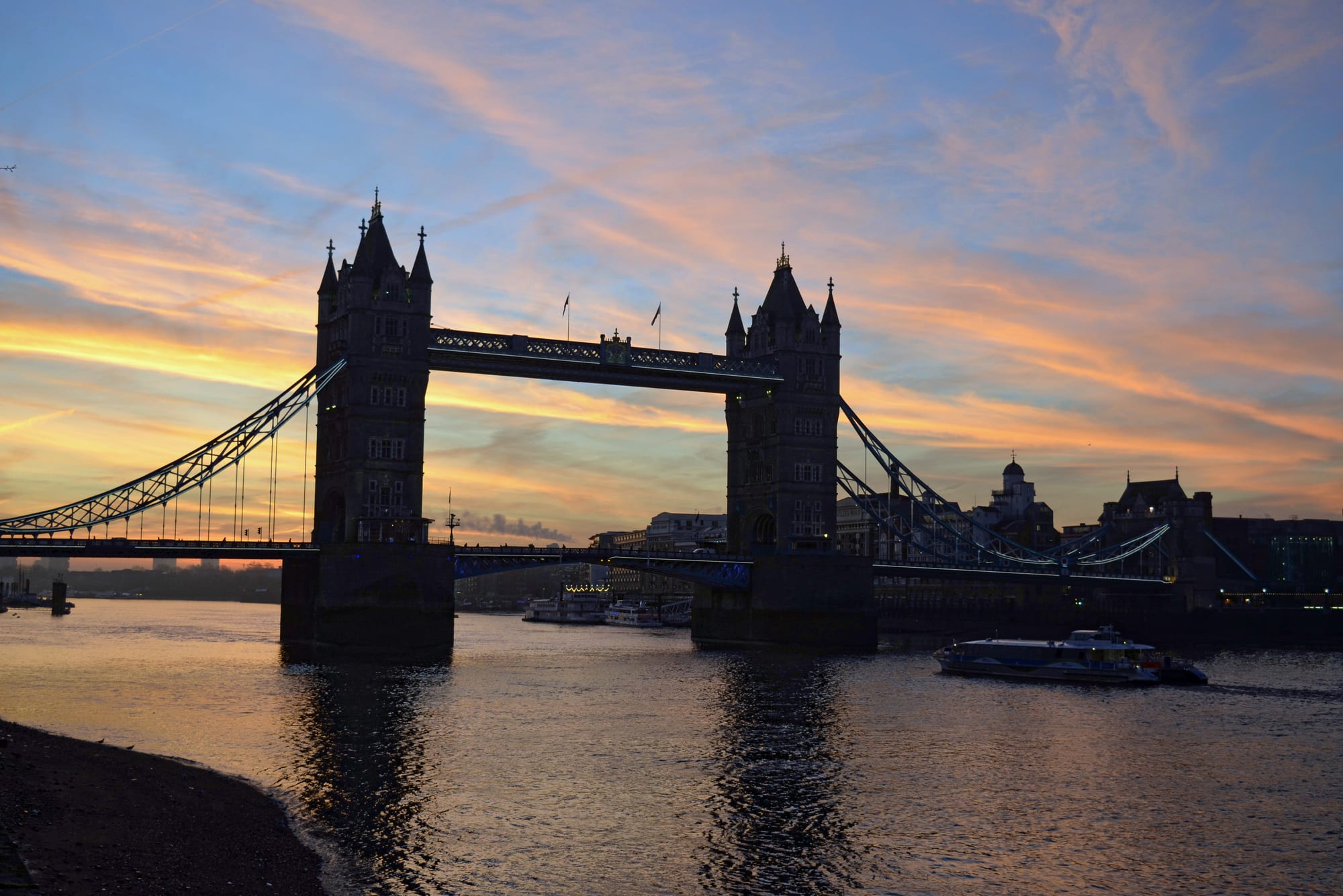 The silhouette of a twin-towered bridge over a calm river, against an orange and blue dawn sky