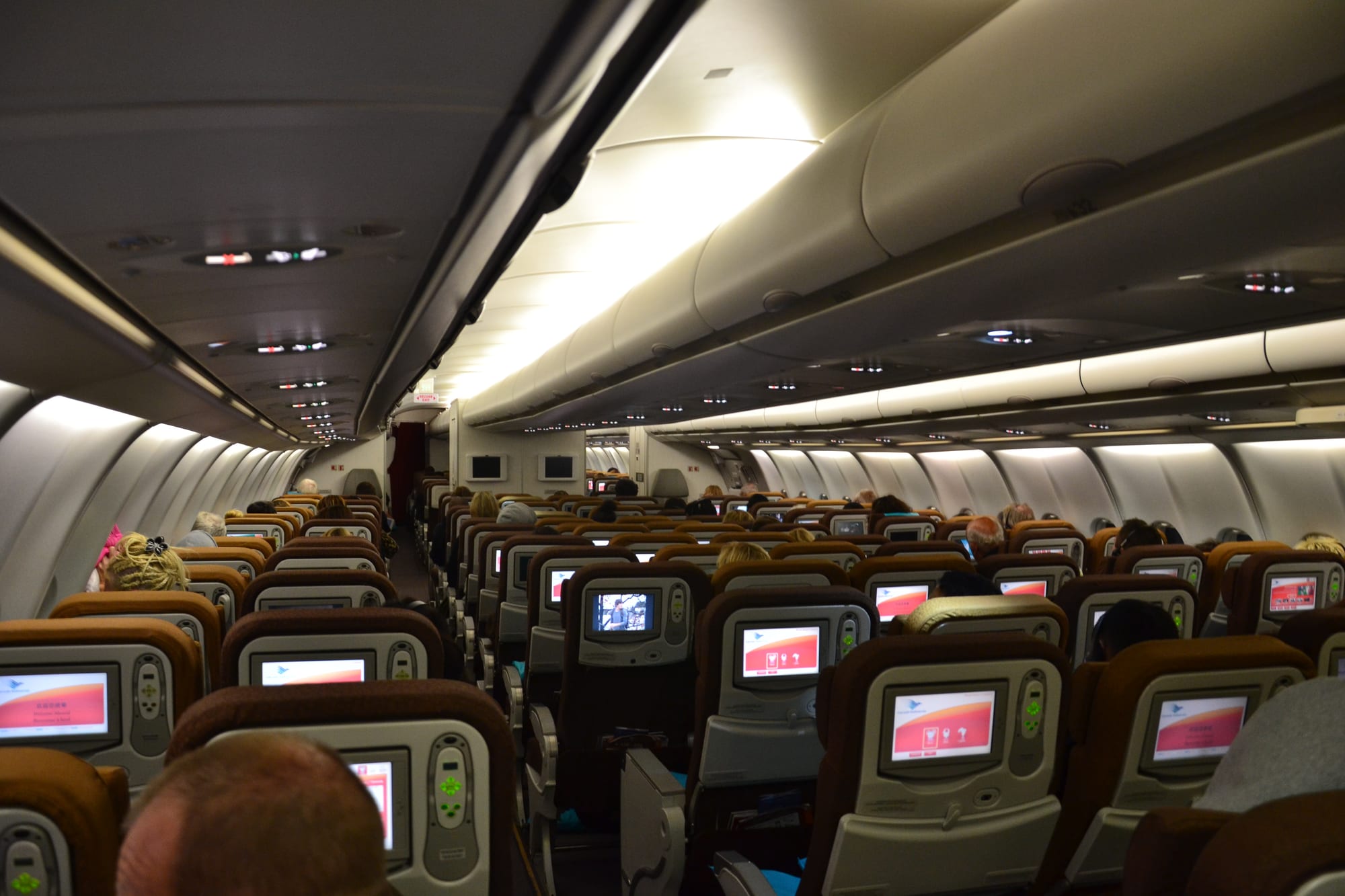 Rows of passengers in a dimly lit economy cabin, each seat back fitted with a small screen