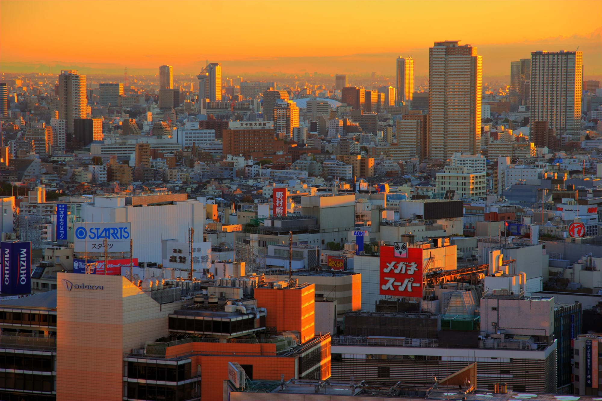 A dense city of office towers and apartment blocks glowing orange under a golden sunrise sky