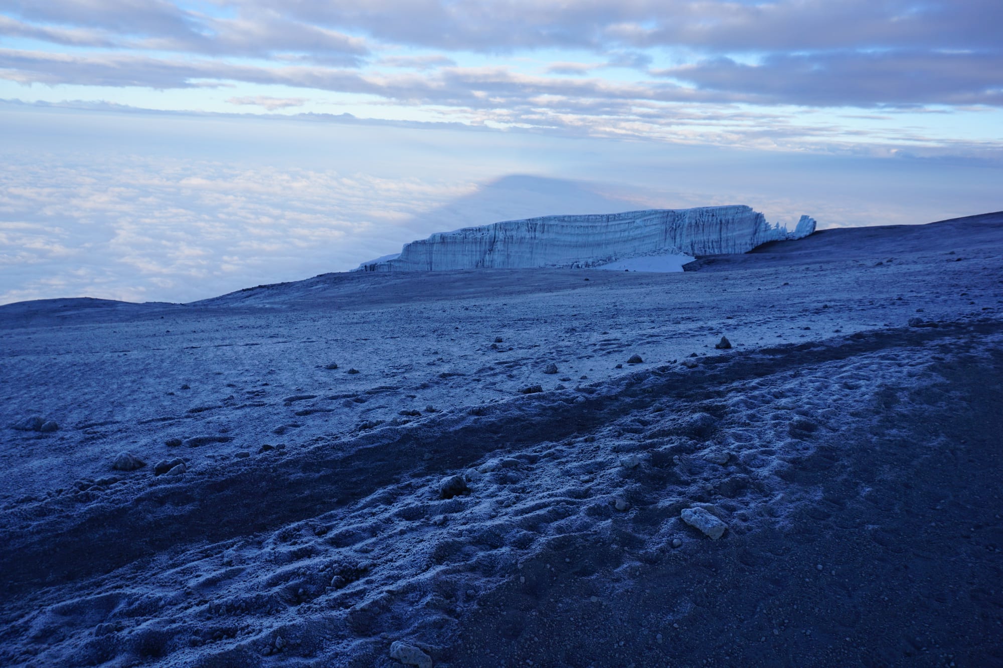 A long wall of glacier ice on a frost-covered summit plateau at dawn, above a sea of cloud