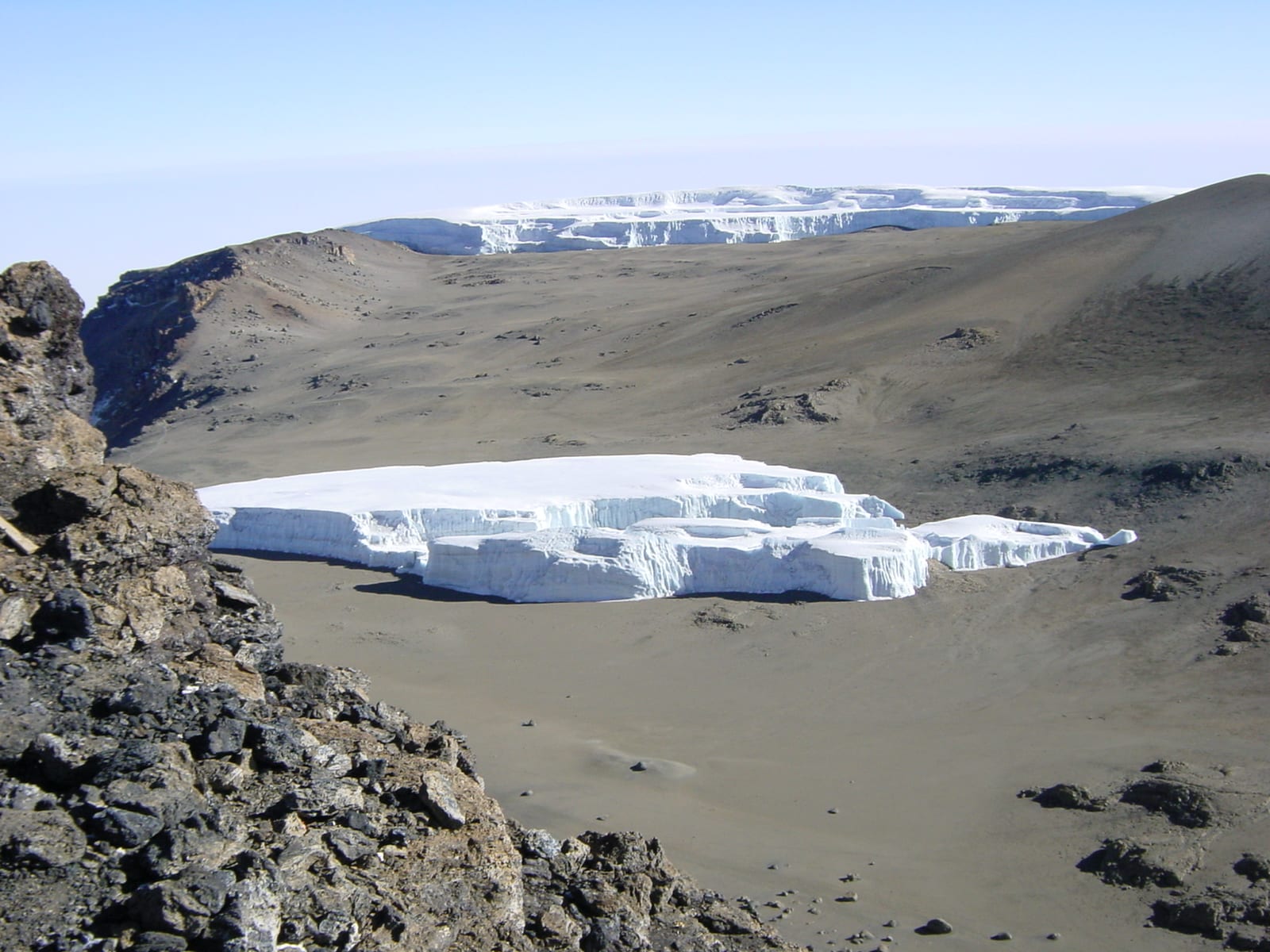 A flat-topped slab of white glacier ice with sheer sides sitting on a bare brown crater floor, with a larger ice field on the rim beyond