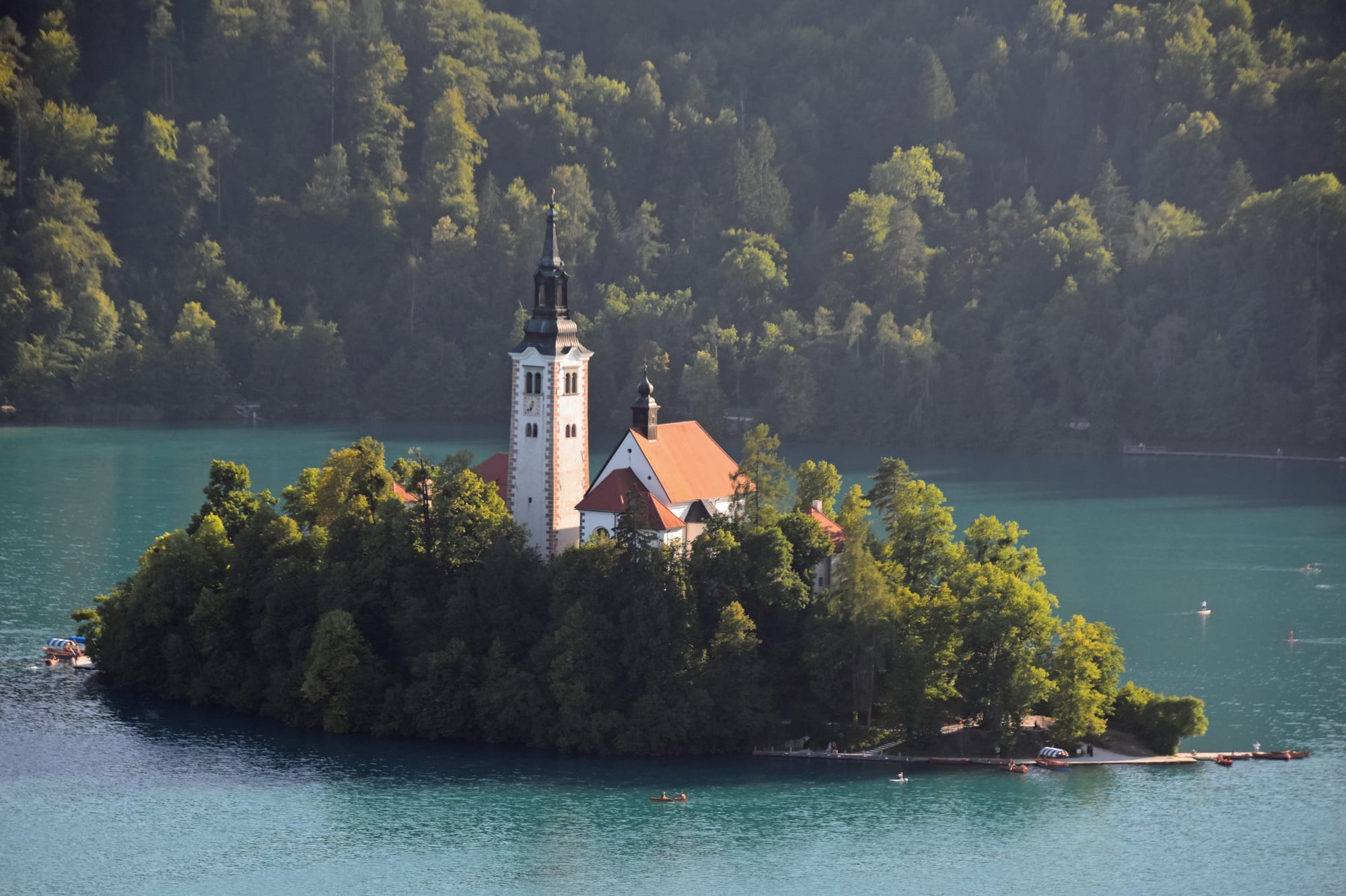 A white church tower with a dark spire and a red-roofed church rising from the trees of a small island in turquoise water