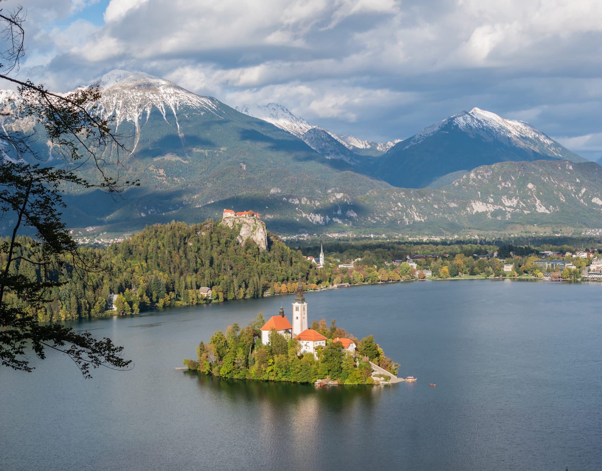 A small wooded island with a white church tower and red roofs in a blue lake, with a castle on a cliff and snow-capped mountains behind
