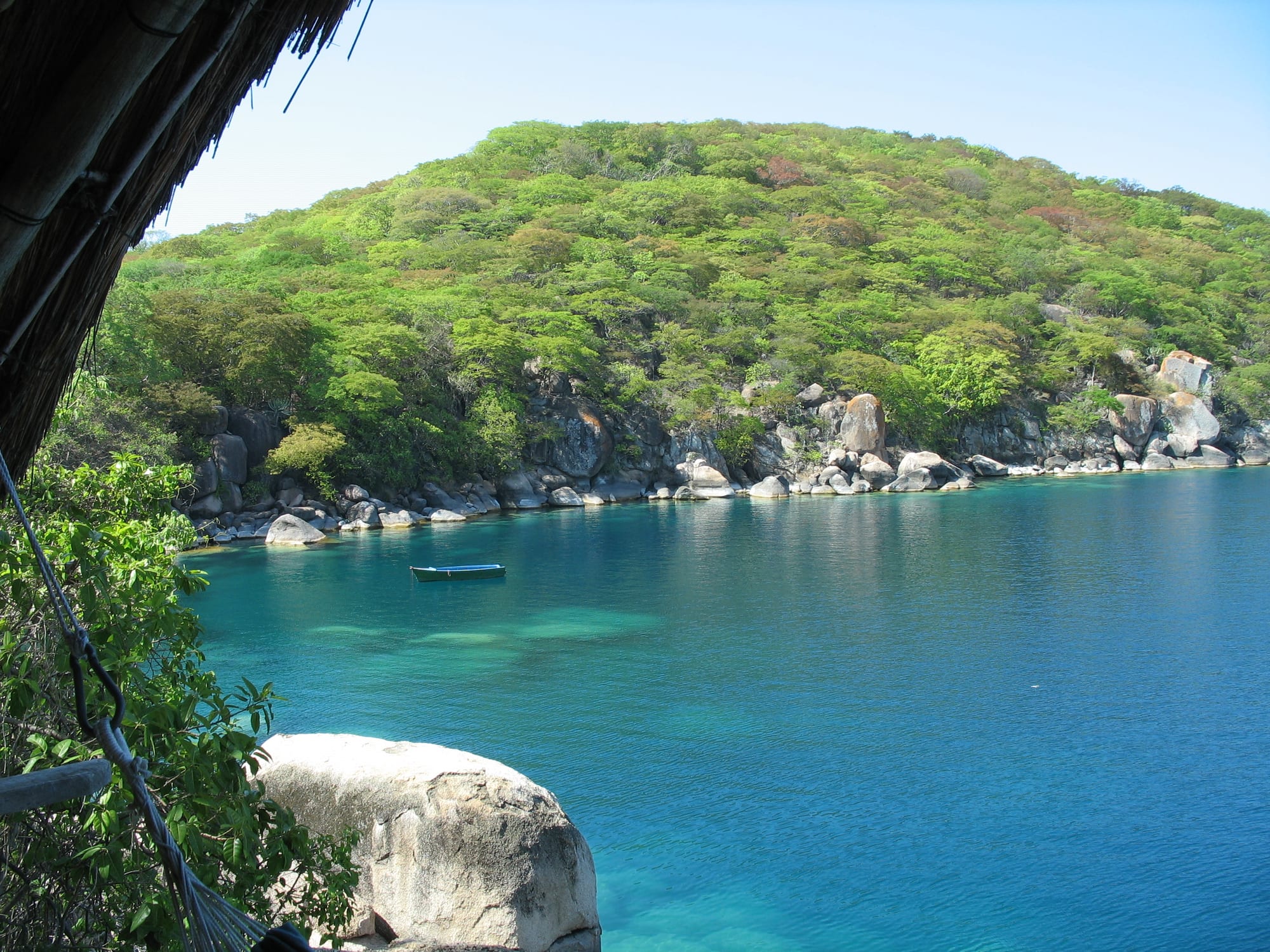 A green, wooded island with a shoreline of pale granite boulders rising from clear turquoise water, with a small boat moored in the bay